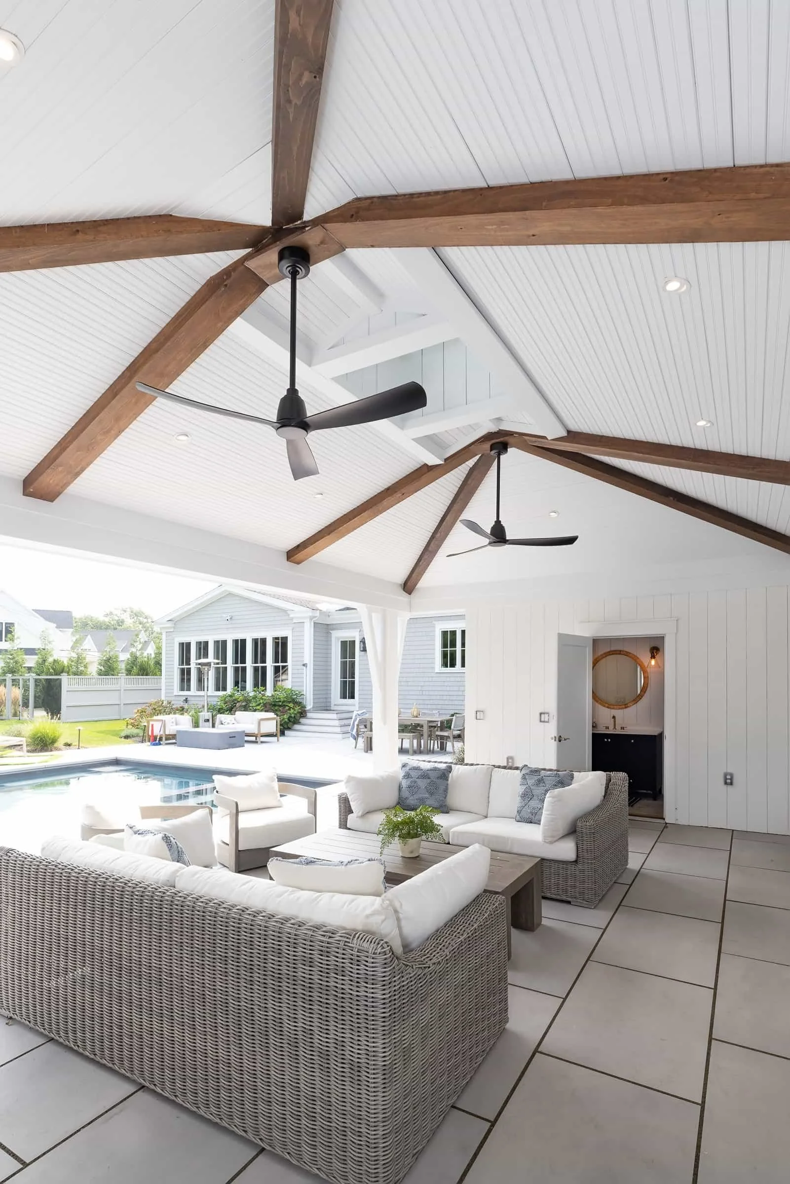 A covered outdoor patio area with white wicker couches and cushions, a wooden coffee table with a potted plant, ceiling fans, and views of a swimming pool and modern backyard with outdoor furniture.