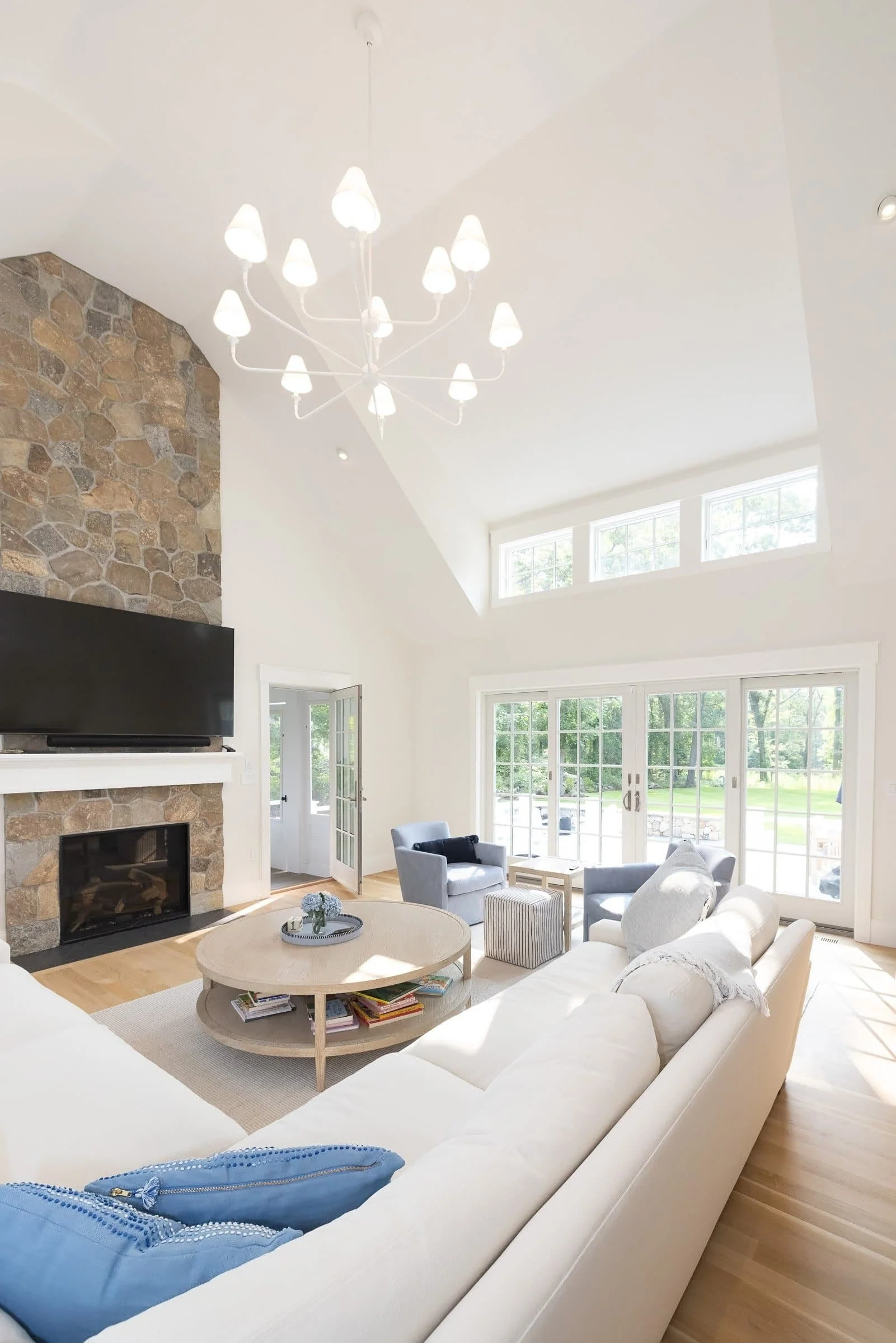 Bright living room with high ceiling, large windows, white walls, stone fireplace, and modern furniture including a white sofa, blue armchairs, and a round coffee table.