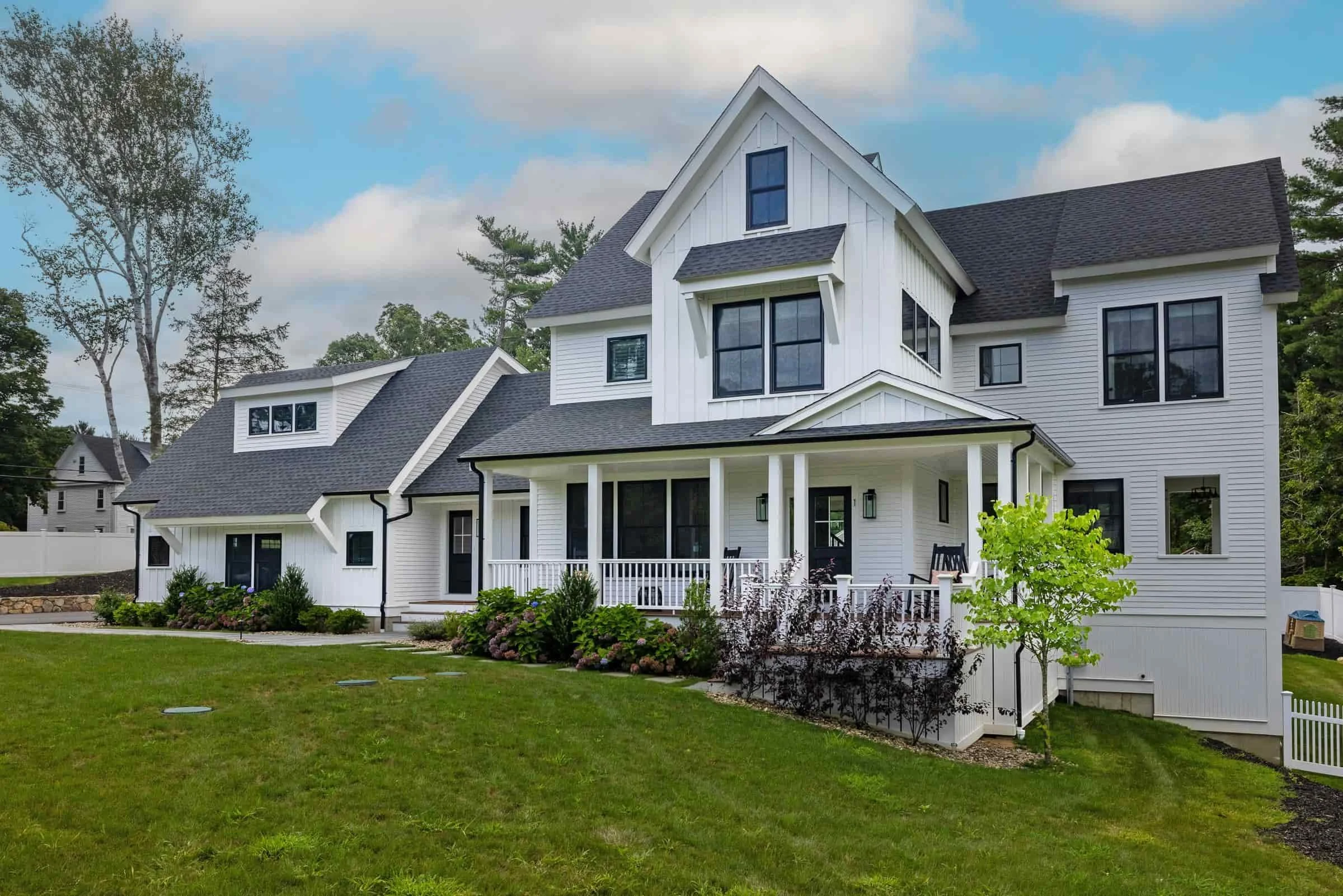 White multi-story house with black roof, front porch with white railing, and landscaping with grass and bushes.