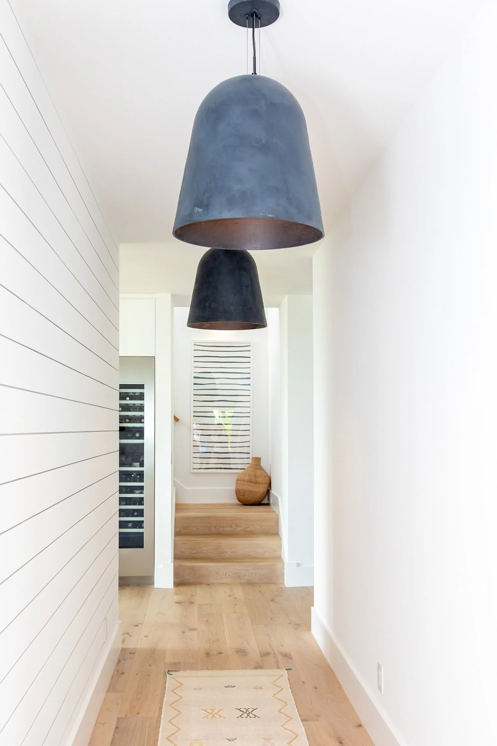 Modern entryway with two black pendant lights hanging from the ceiling, a wooden floor, a small rug, a staircase with a woven basket, and a window with striped blinds.