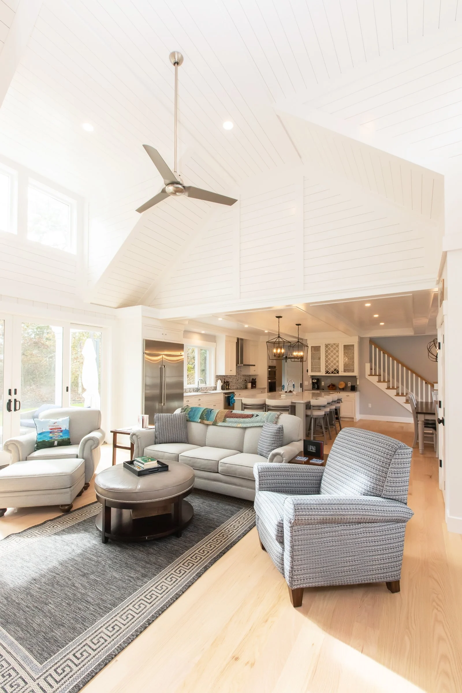 Bright open-concept living room with white walls, high vaulted ceiling with wood paneling, ceiling fan, large windows, and a view of the kitchen and staircase.