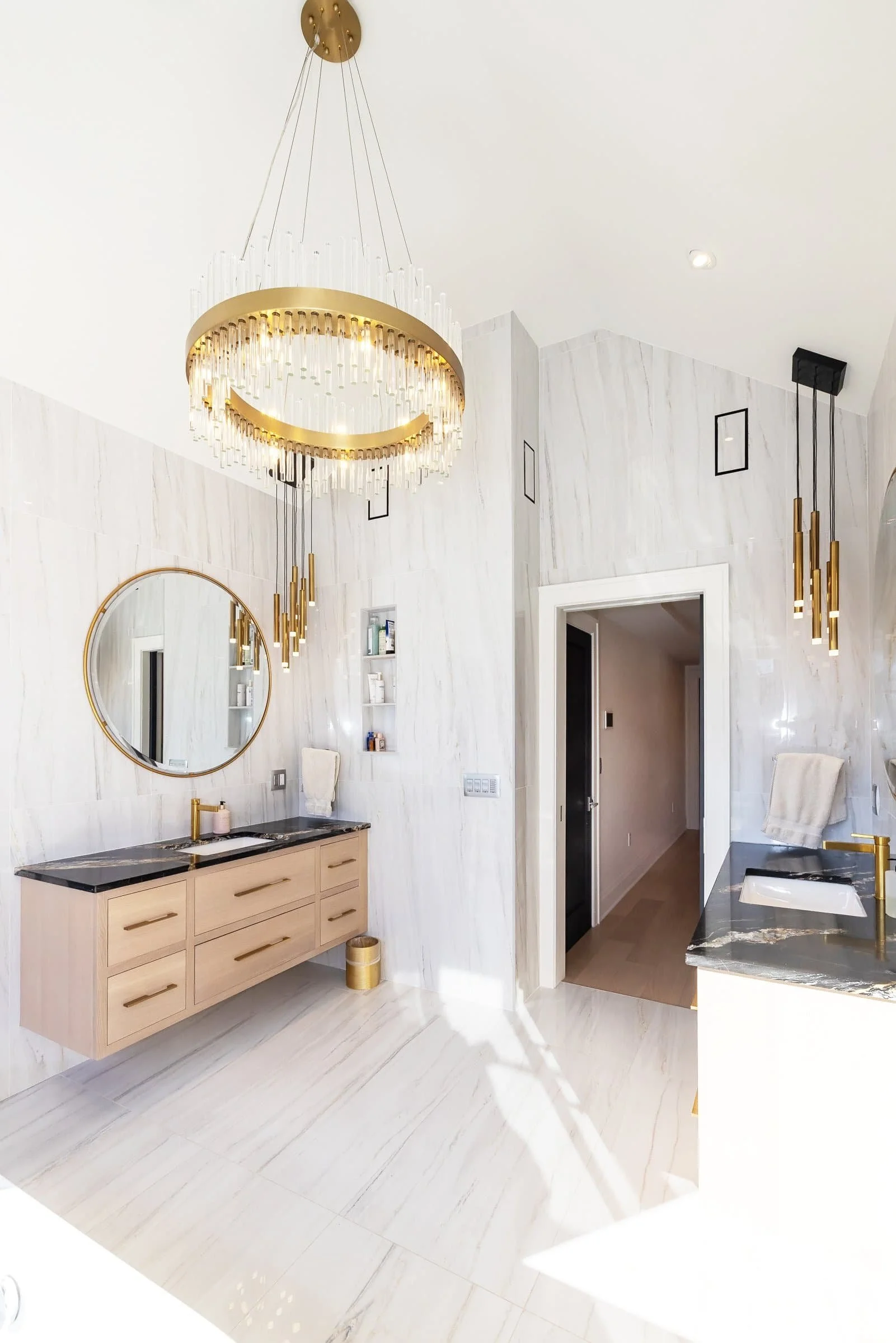 Modern bathroom with white marble walls and floors, a large circular chandelier, gold and black accents, a floating vanity with black marble countertop, a round mirror, and a black and white sink.