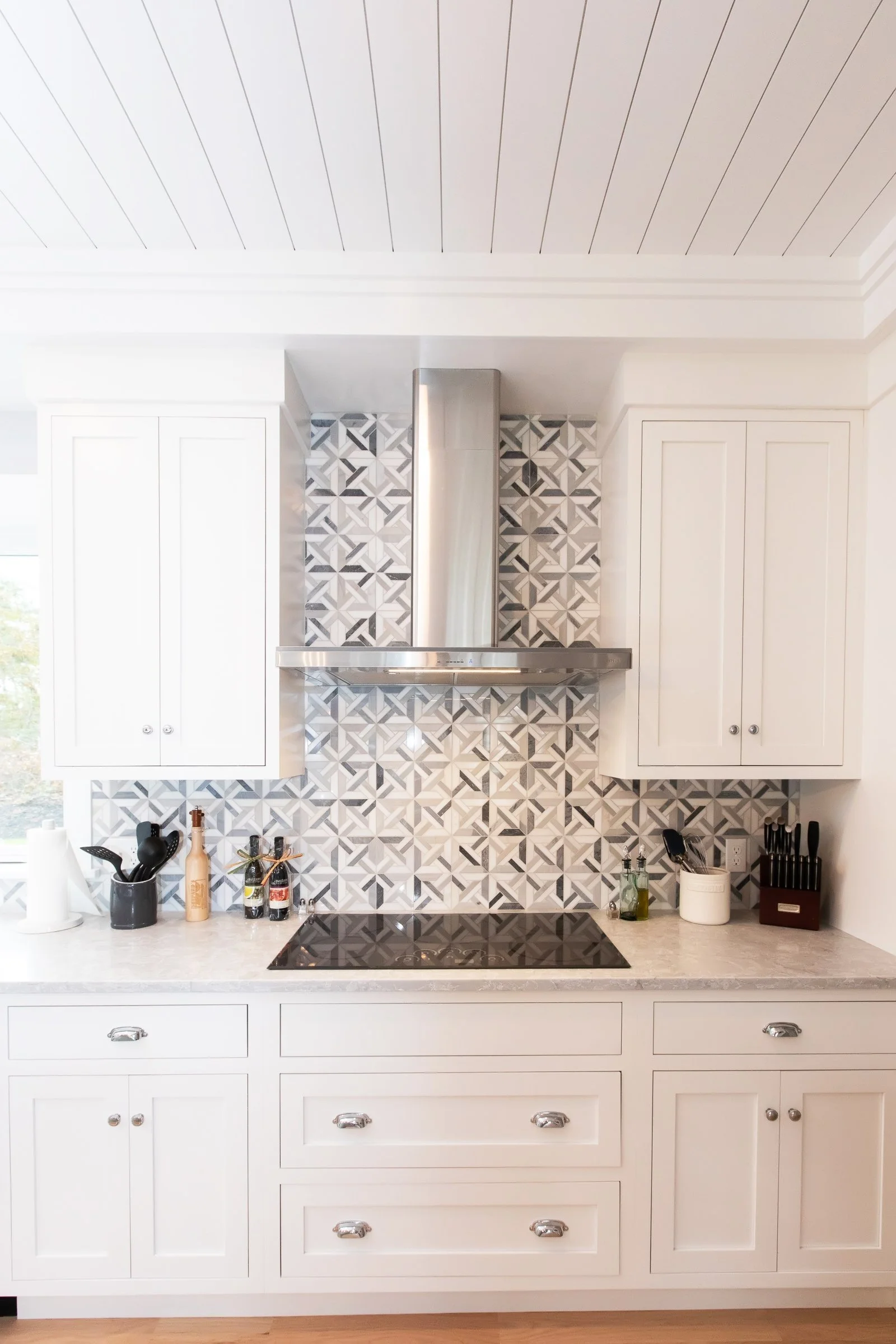 White kitchen with patterned tile backsplash, black stove top, stainless steel range hood, and white cabinets.