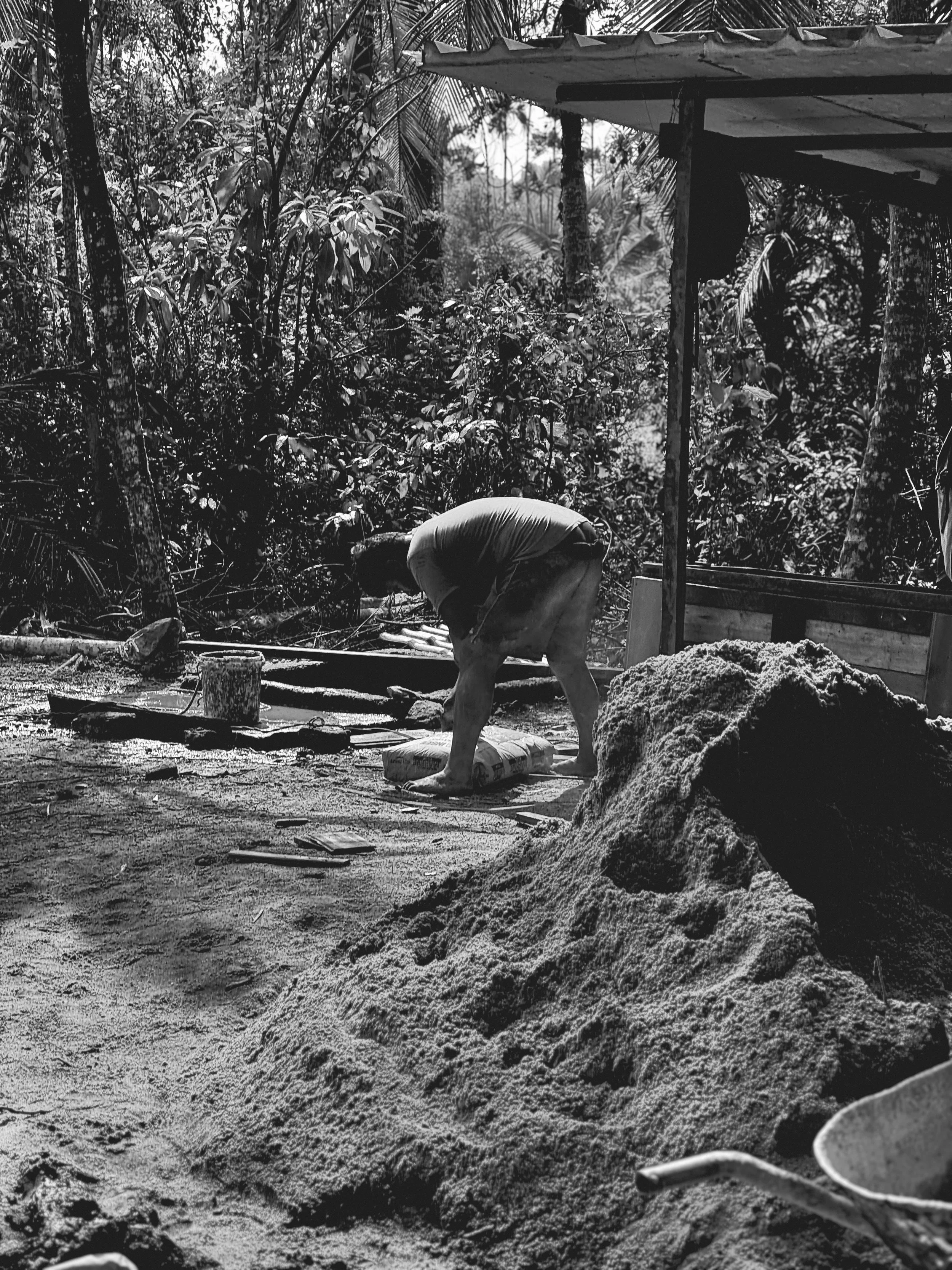 A person working outdoors in a tropical forest setting, bending over a task with a pile of sandy material nearby.