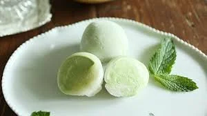 Three round green mochi ice creams and a mint leaf on a white plate.