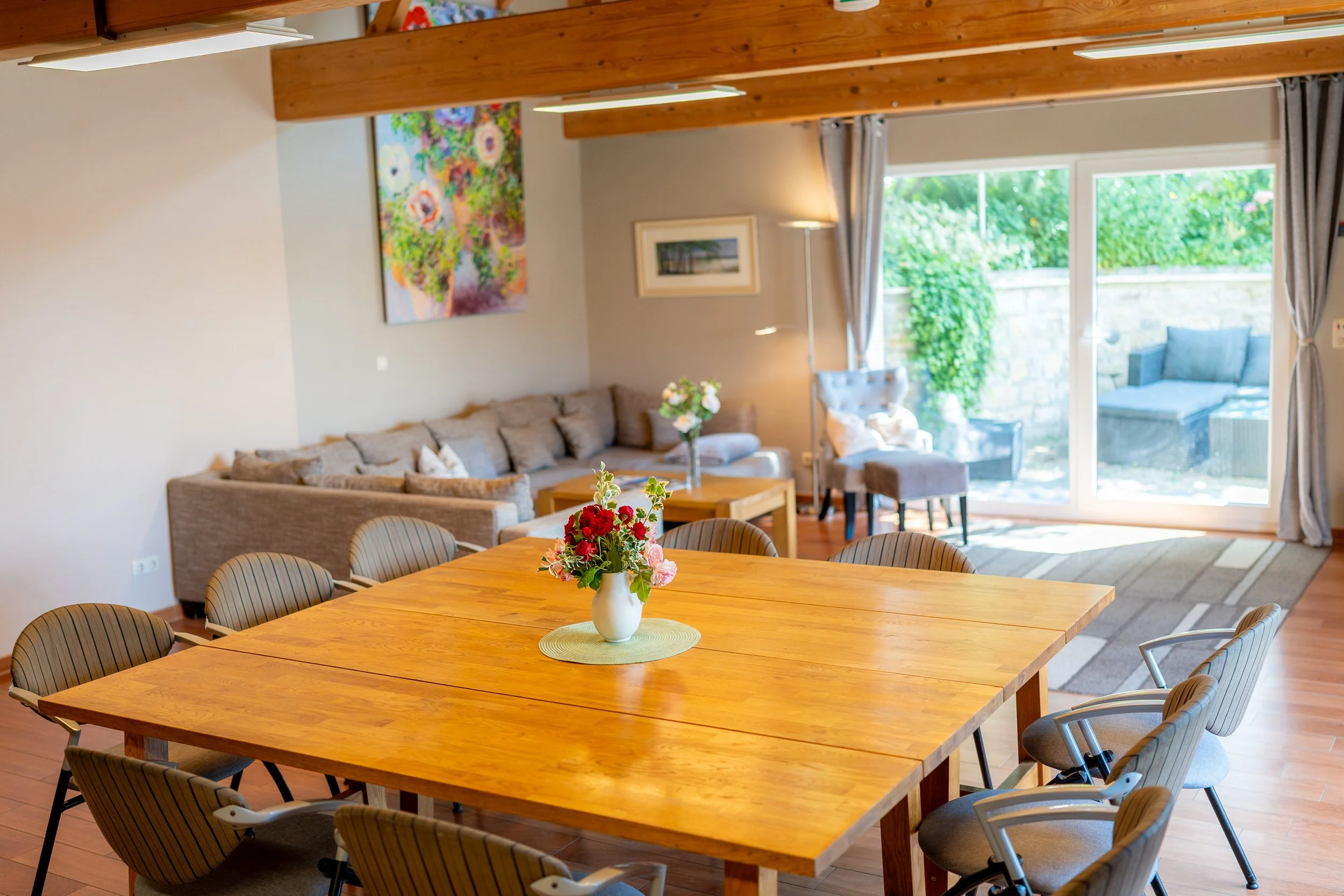 Moderner Wohn- und Essbereich mit Holzdecke, großen Fenstern mit Vorhängen, Couch, Armchairs, Blumenvasen auf Tischen und Wandbildern.