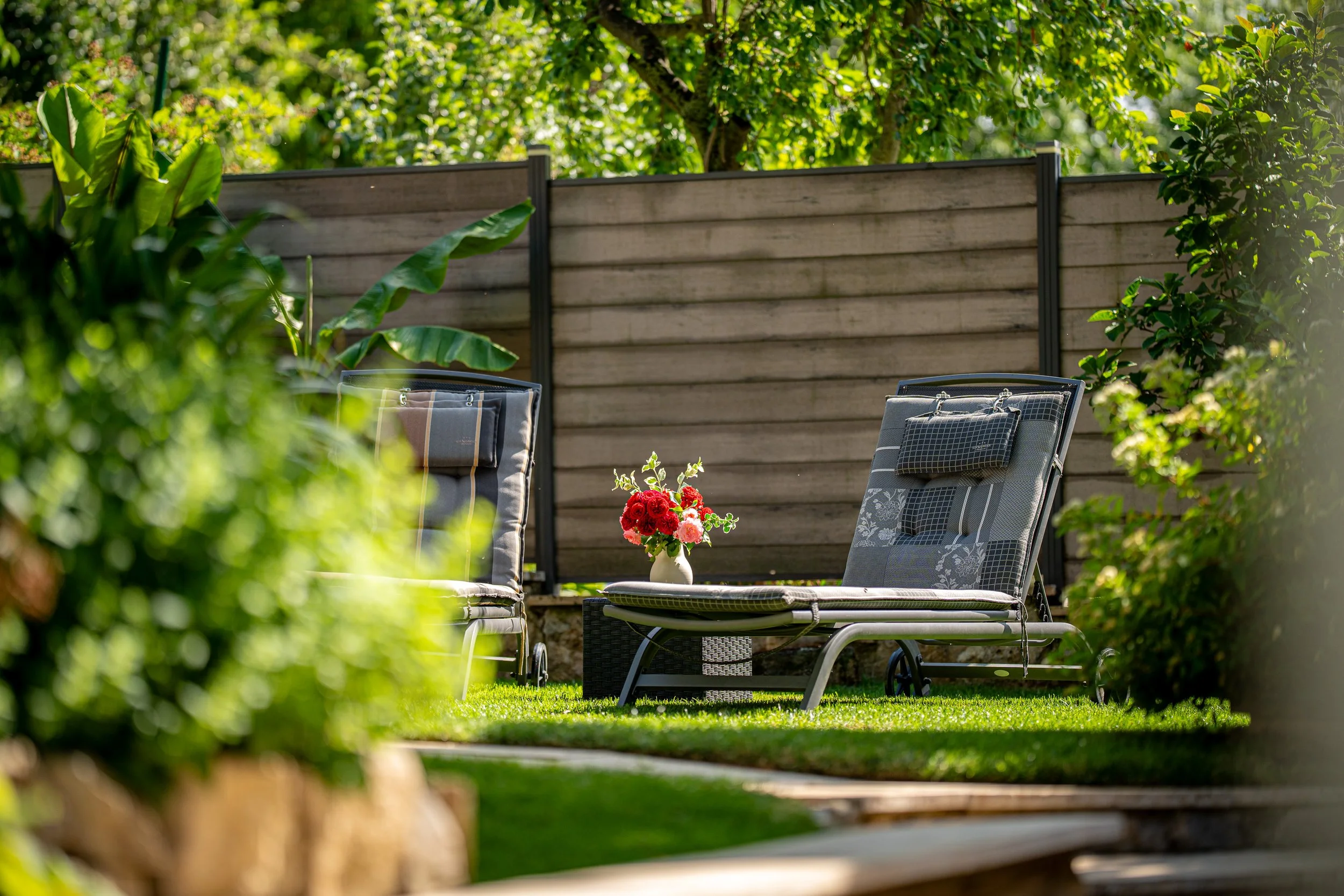 Garten mit Liegestuhl, einem kleinen Tisch mit einer Vase voller rote und rosa Blumen, umgeben von grünem Pflanzen und Bäumen, eingezäunt mit Holzlattenzaun.