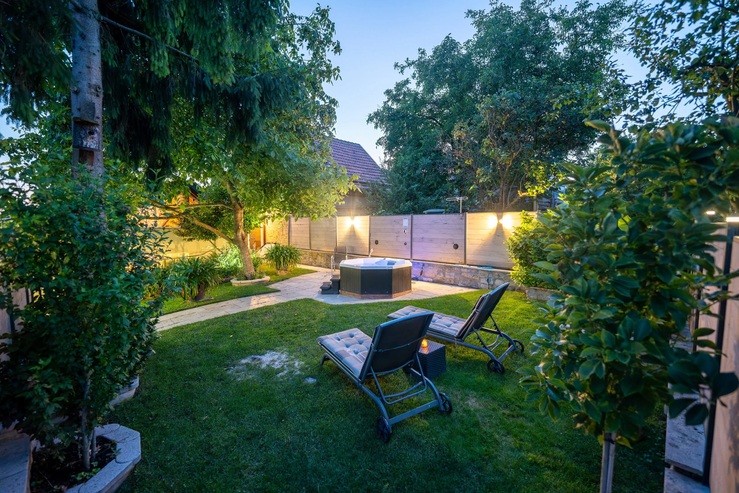 Garten mit Rasen, Liegestühlen, einem beheizten Whirlpool, Bäumen und einer Holzsichtschutzwand bei Abenddämmerung.