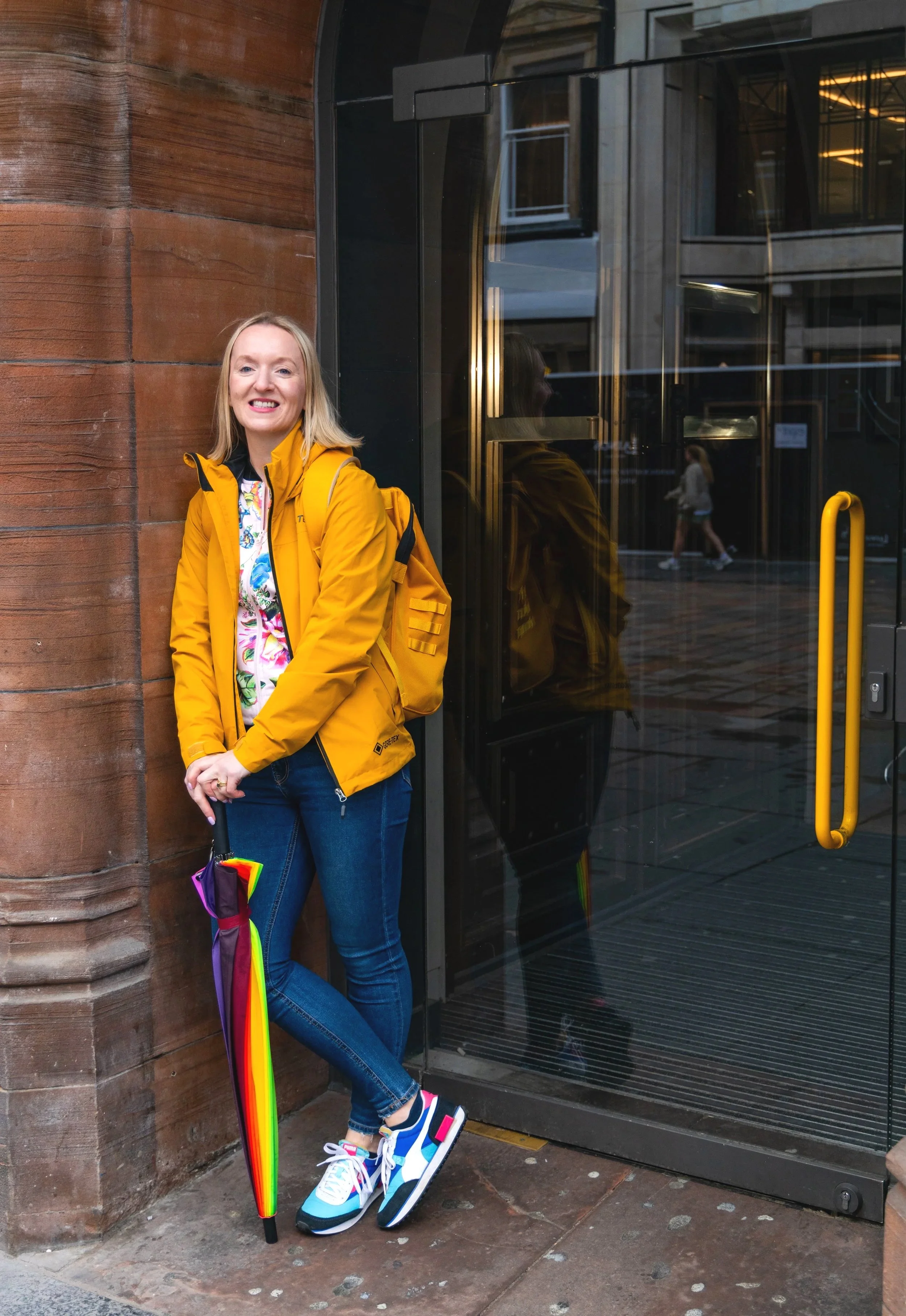 A woman in a yellow jacket standing next to a glass door on a city street, holding a rainbow umbrella and wearing colorful sneakers.