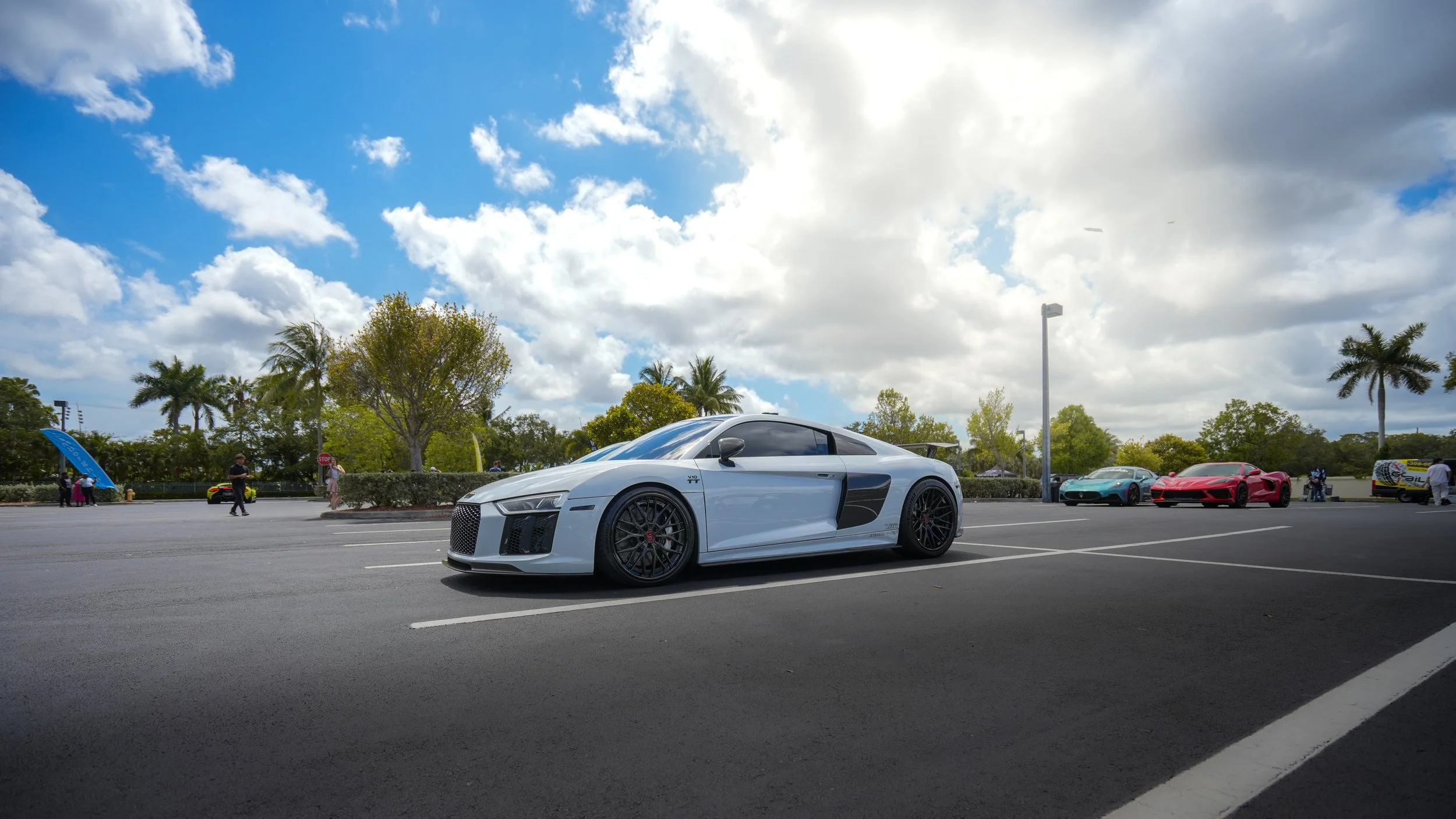 White Audi R8 sports car parked in a parking lot with palm trees, colorful sports cars, and a partly cloudy sky in the background.
