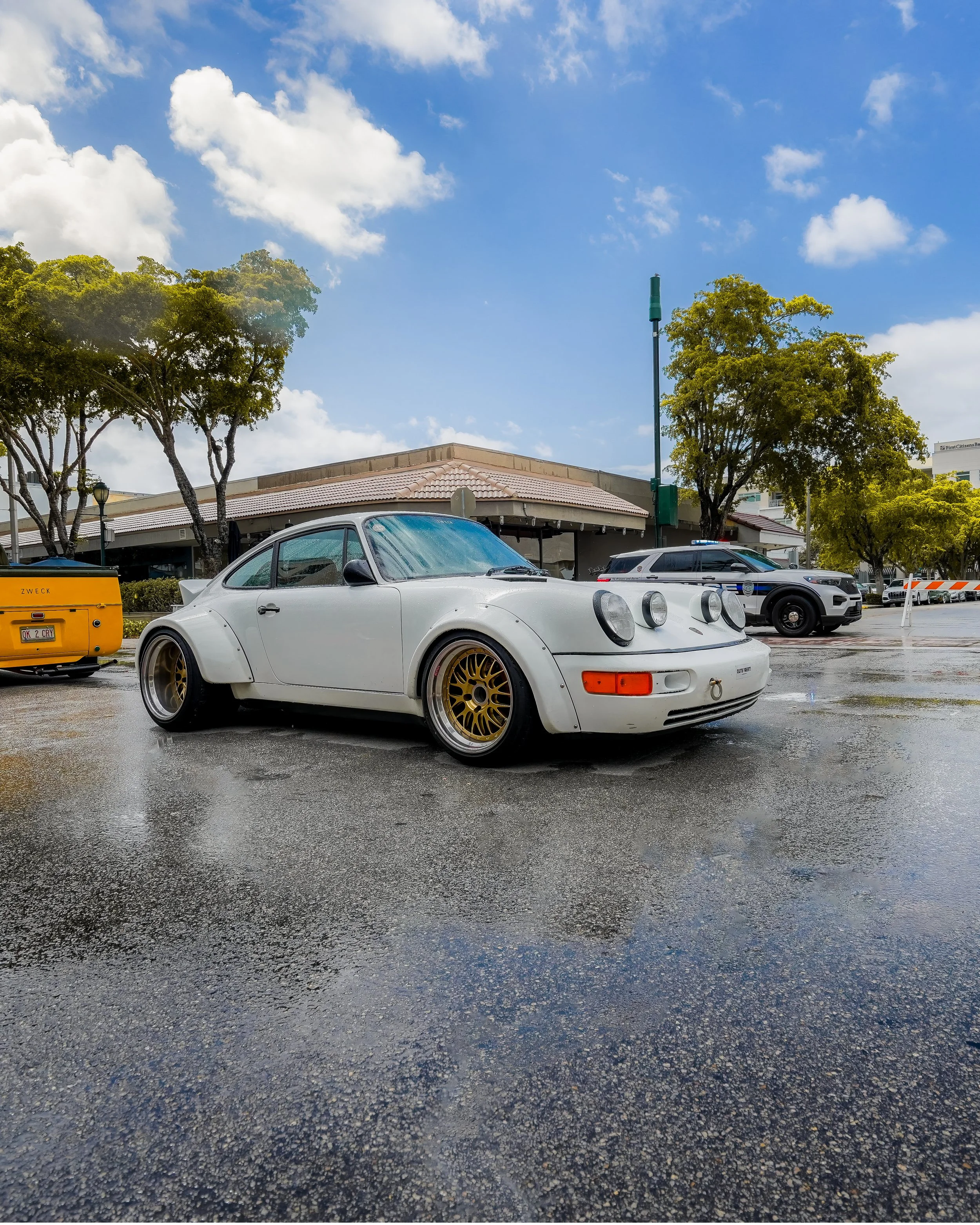 White vintage Porsche 911 parked on wet pavement, with a yellow car and police vehicle in the background under partly cloudy sky.