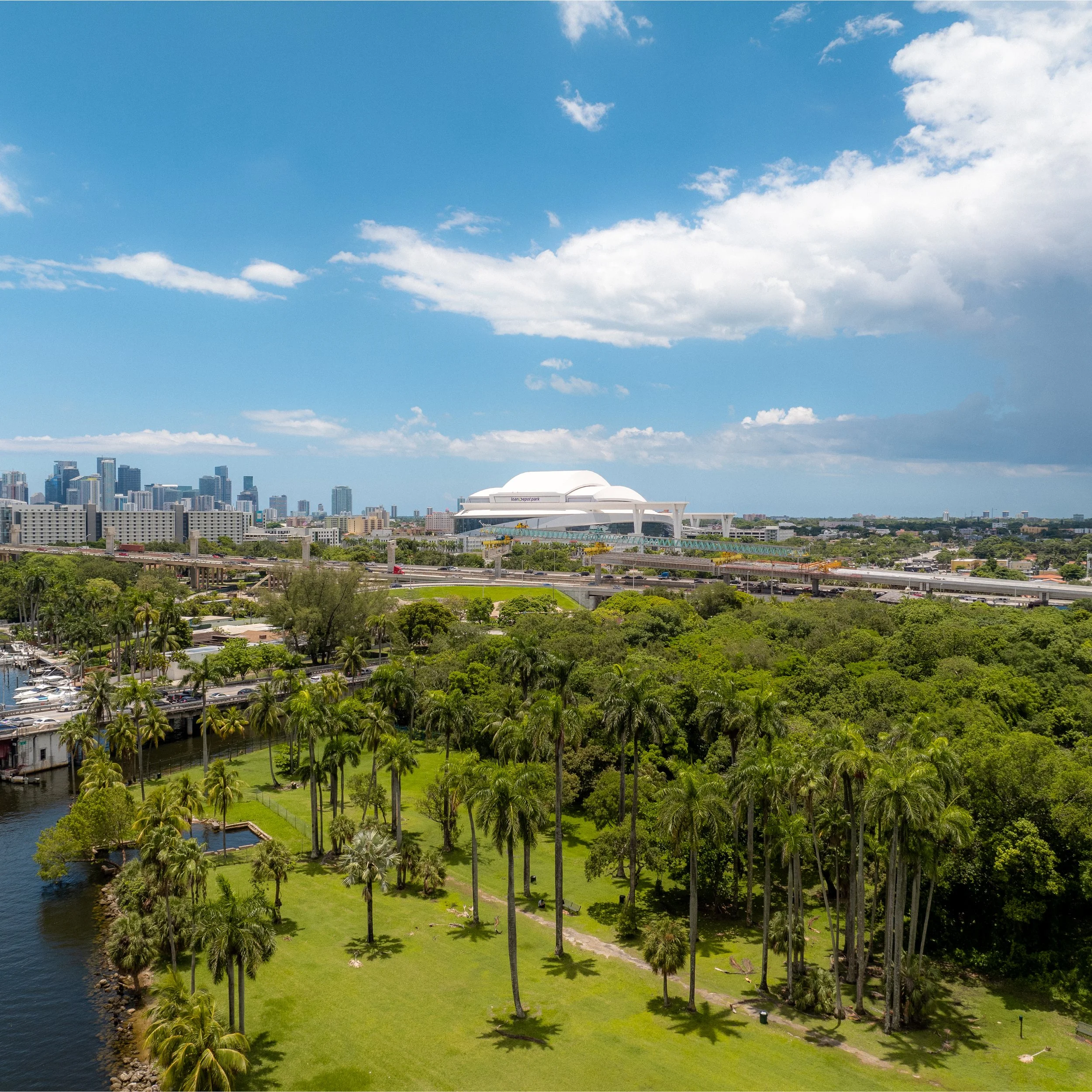 City skyline with a white modern building, green park with palm trees, water, and highways under a partly cloudy sky.