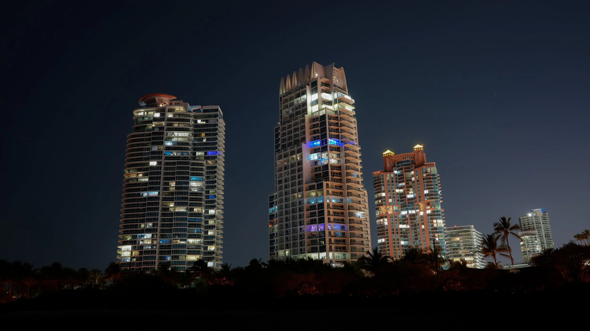Night view of tall, illuminated skyscrapers with modern architecture, set against a dark sky and palm trees in the foreground.