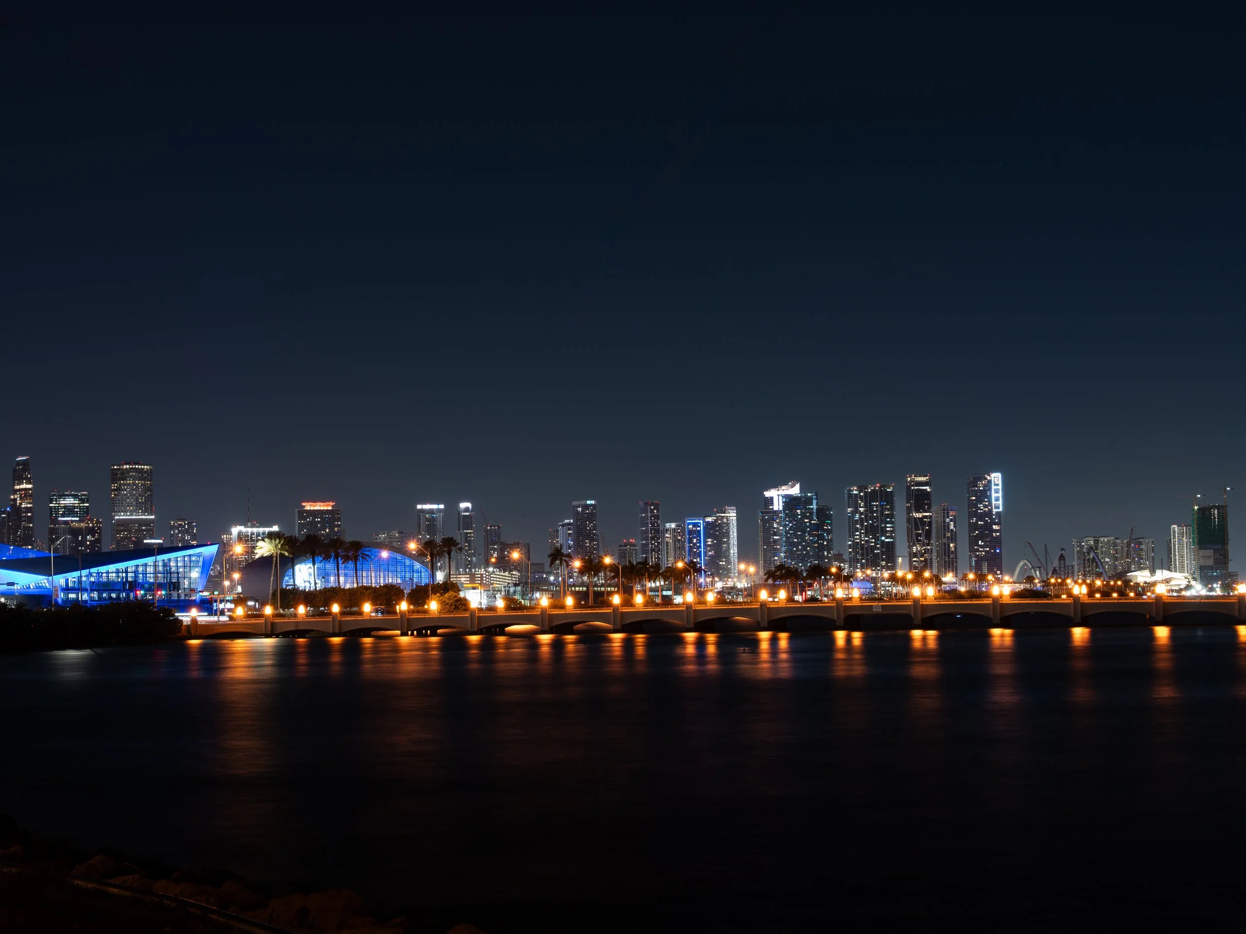 Nighttime city skyline with tall illuminated buildings, waterfront, and reflections on the water.