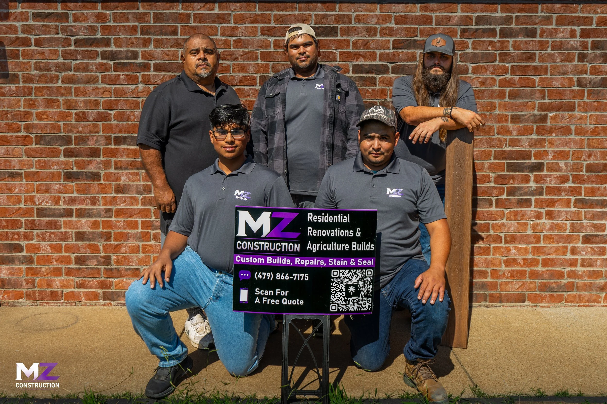 Five men standing in front of a brick wall, holding a sign advertising construction services. The sign includes a phone number, QR code, and text about custom builds, repairs, stain, and seal. Four men are wearing gray shirts with a logo, and two are kneeling while three are standing behind them.