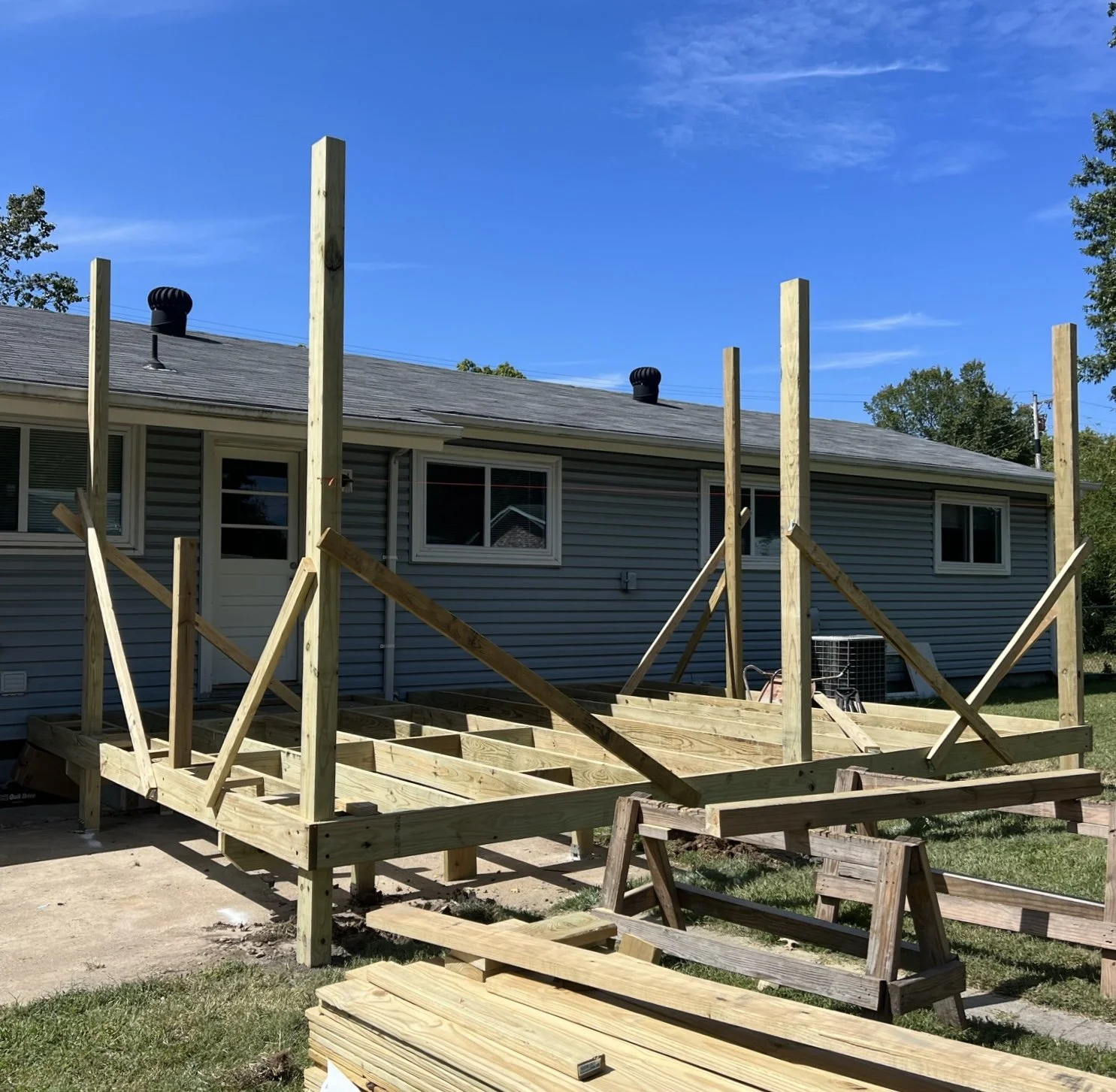 In the process of building a wooden deck attached to a house, with wooden beams and supports in place, and construction tools on the ground, under a clear blue sky.