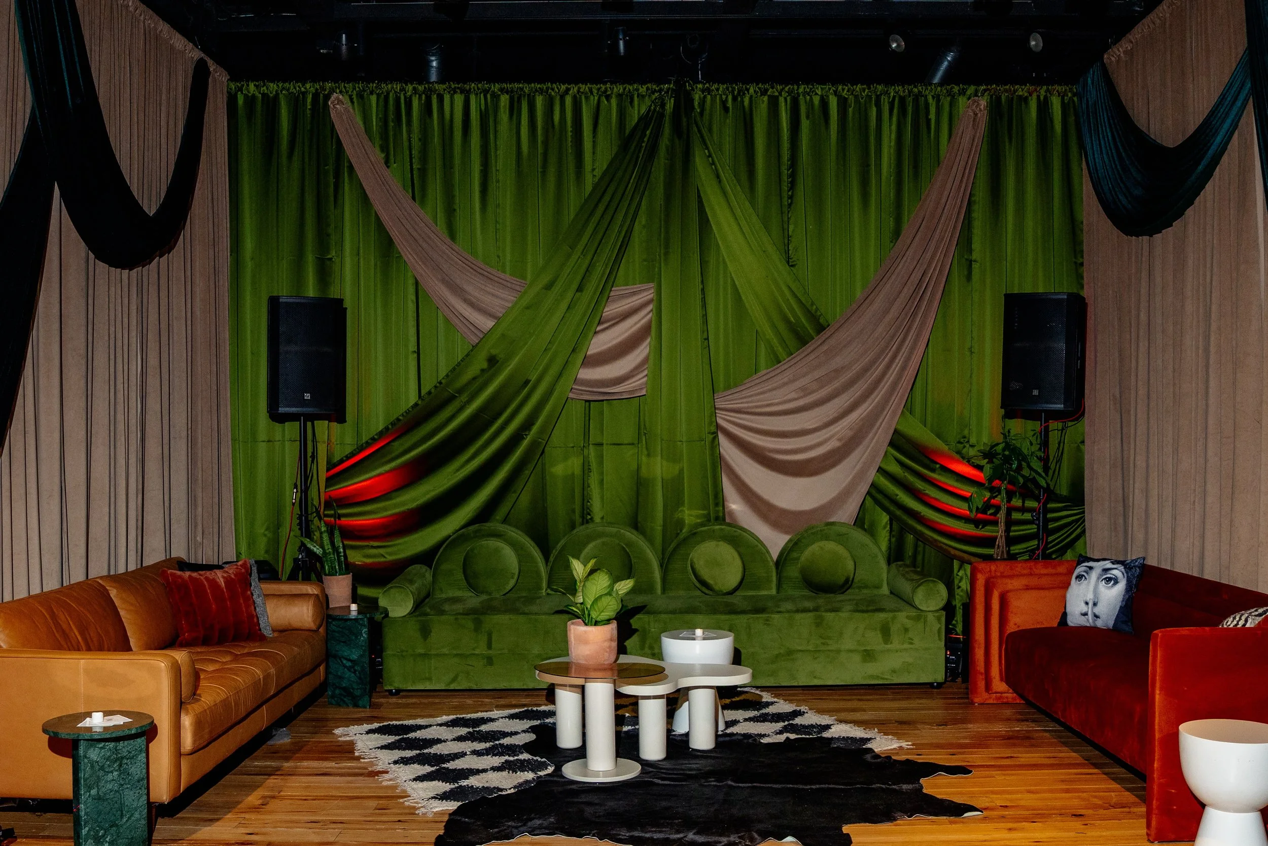 Indoor lounge with a green velvet sofa, orange and red velvet couches, a black and white patterned rug, and a backdrop of green and beige drapes with colorful fabric accents. There are two large black speakers on stands and some plants and decorative pillows.