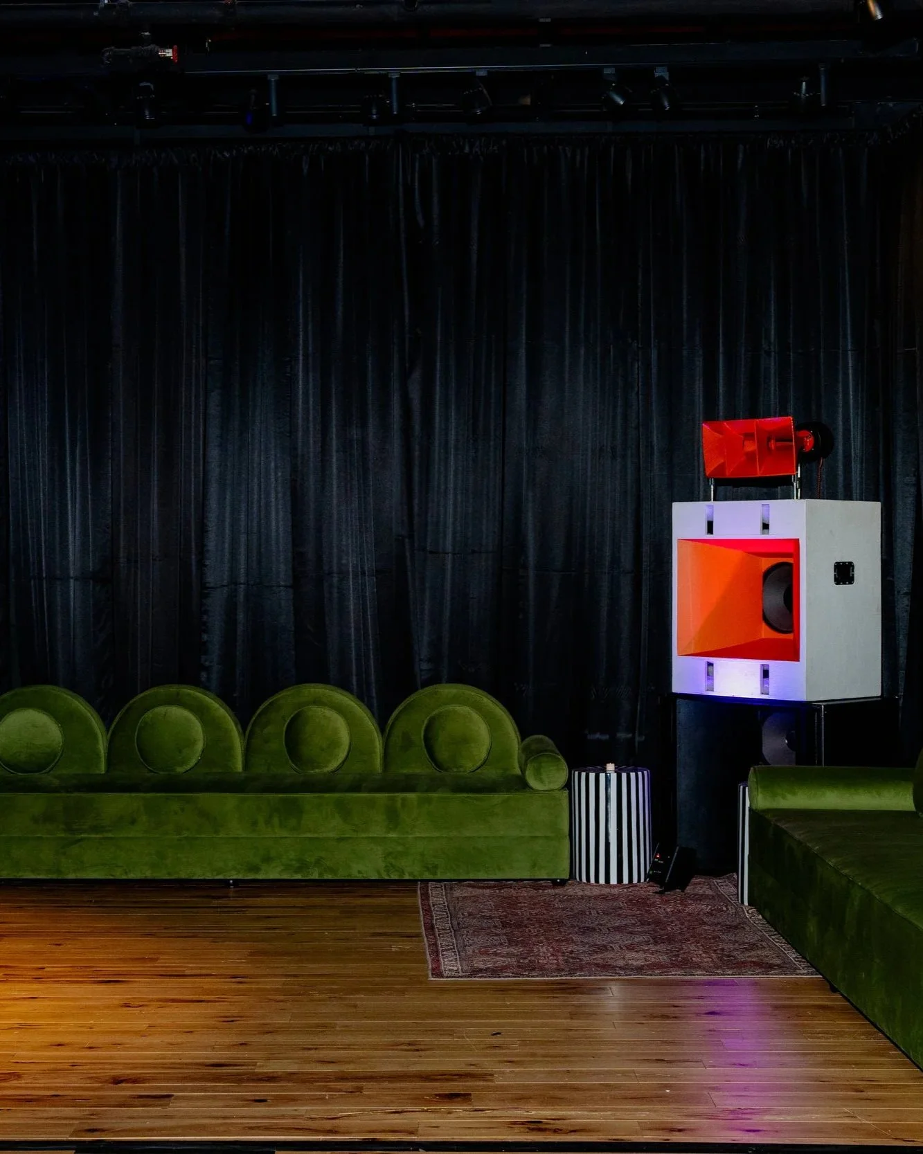 Stage with black curtains, green velvet seating, and a speaker system with a red speaker on white box, and a striped cylindrical container, on a wooden floor.