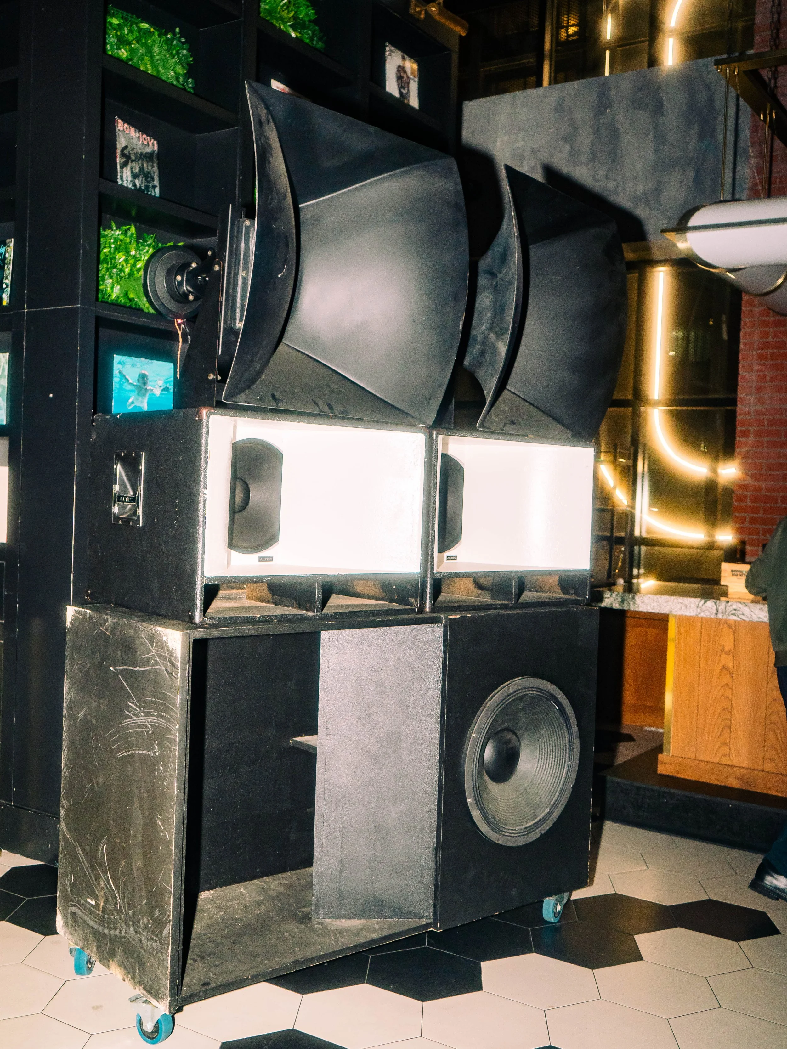 Stacked large black speakers with tweeters and woofers, placed on a black cart with wheels, inside a modern restaurant or bar with black shelving, decorative lighting, and a counter with a marble top.