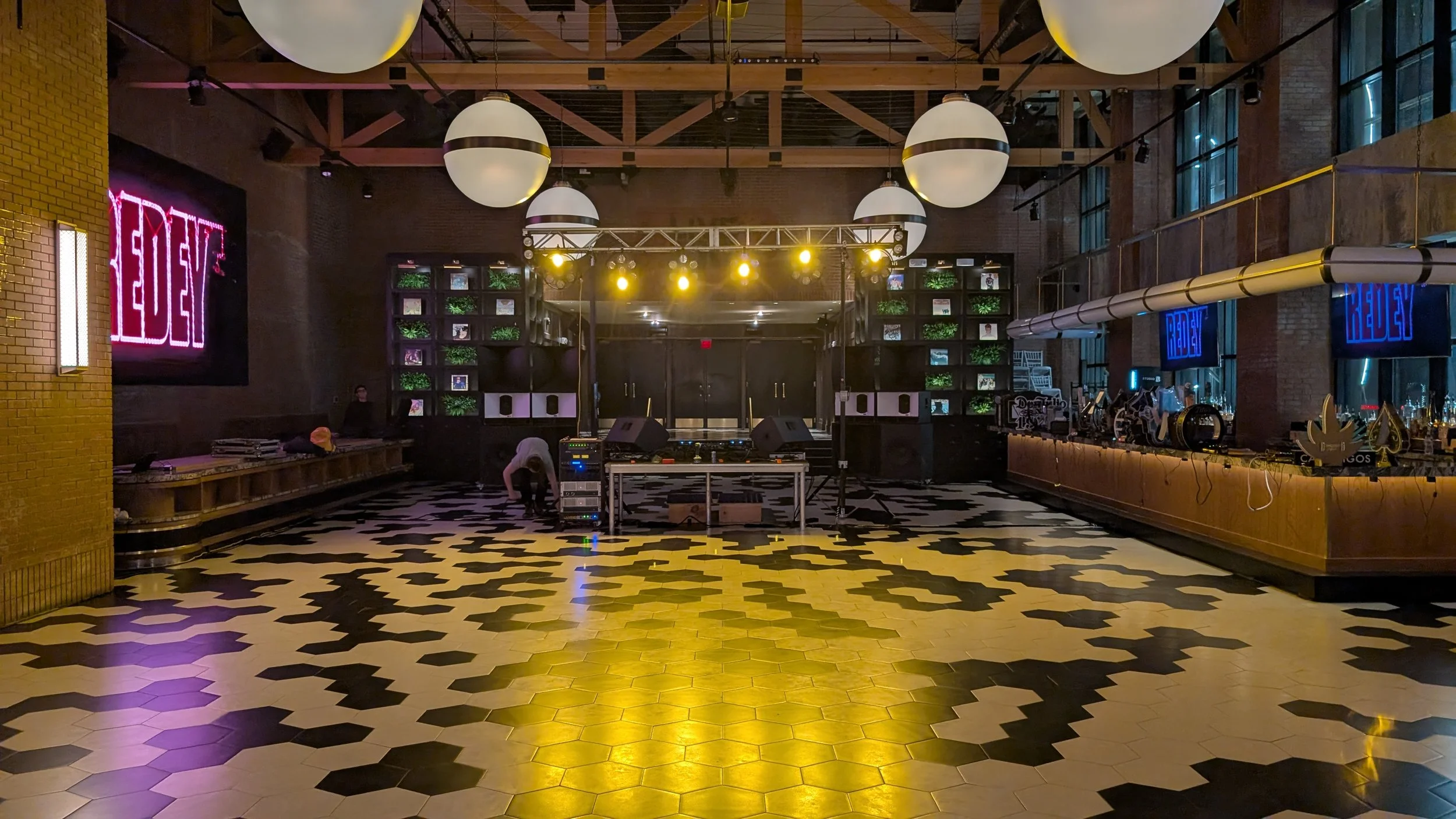 An empty nightclub dance floor with black and white patterned tiles, large hanging white spherical lights, and a DJ booth at the back with sound equipment. Neon sign on the left wall reads 'REVEL', and there are blue digital screens on the right wall