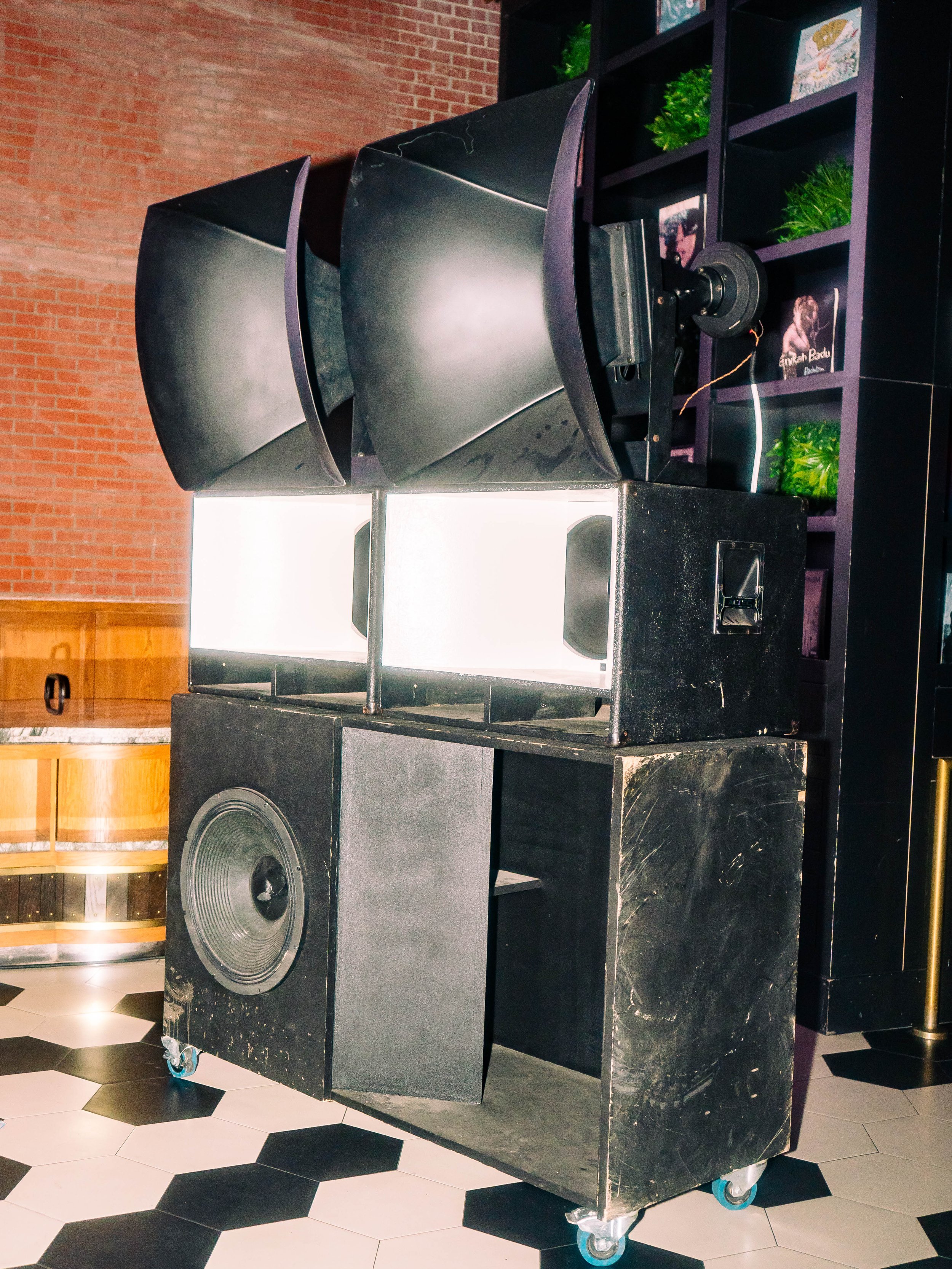A set of large speakers with some modifications on a black cart with wheels, placed on black and white tiled flooring. Photos are visible in the background on purple shelves.