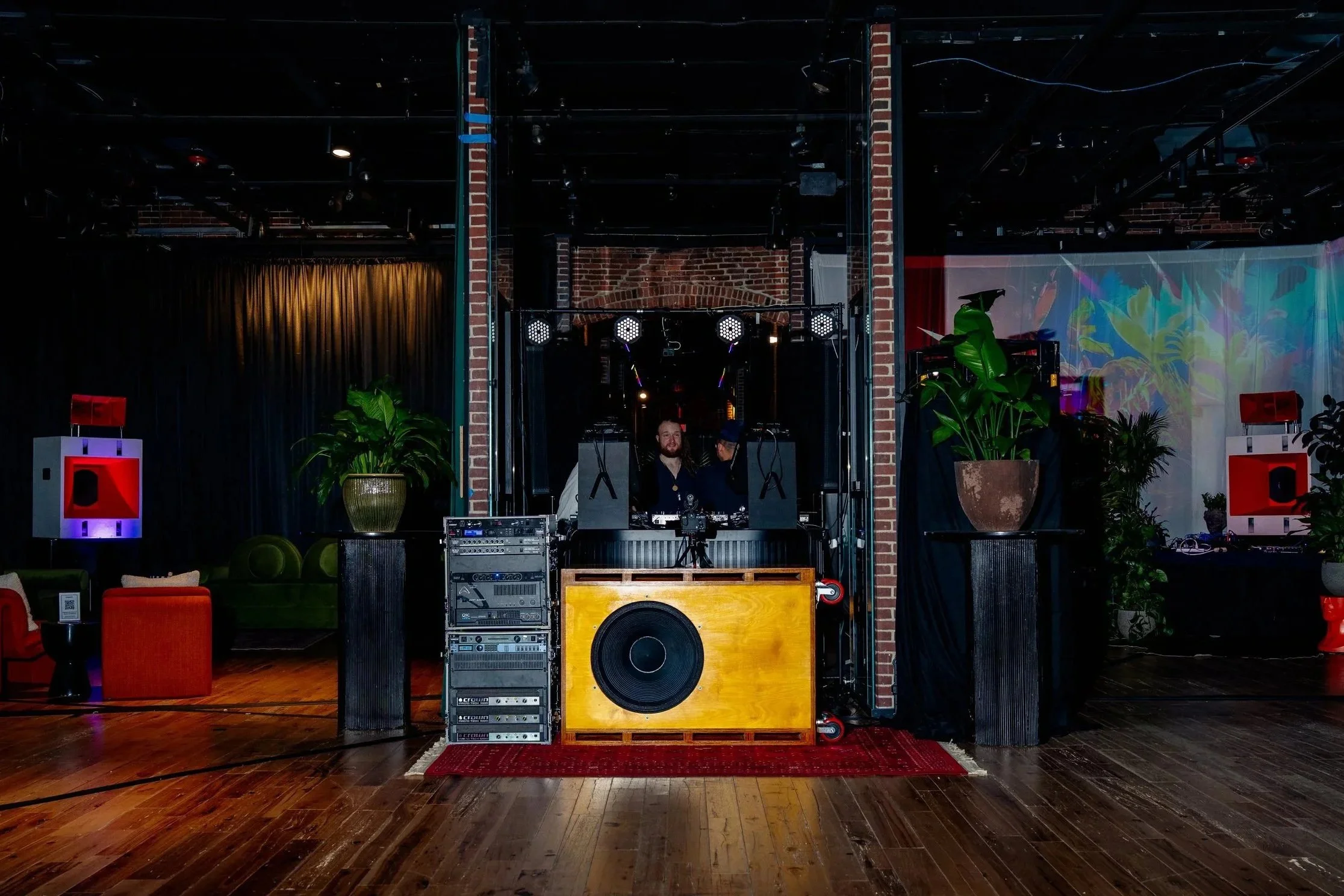 A DJ setup with equipment, speakers, and two people behind it at an indoor venue with seating areas and plants.