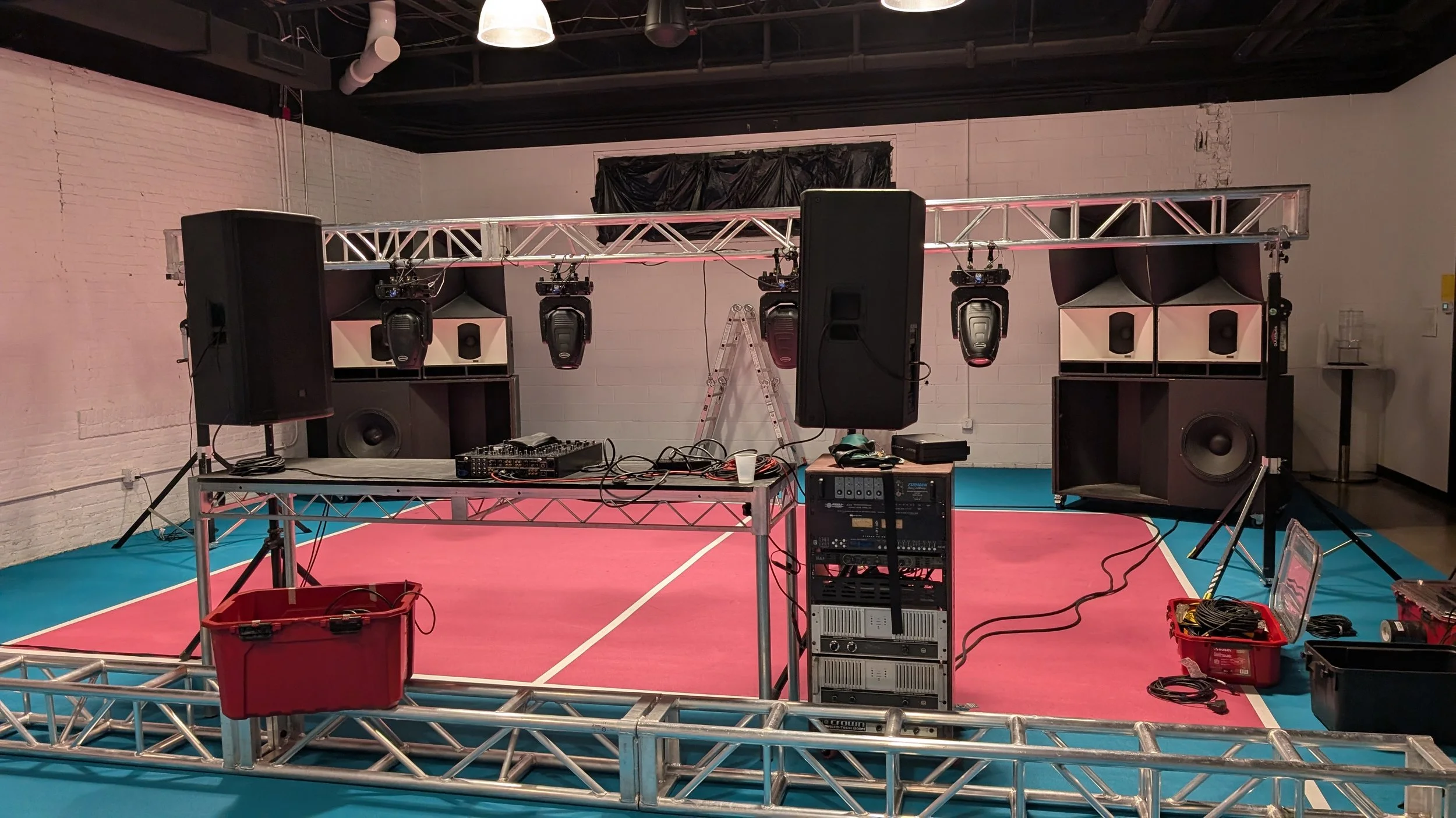 DJ booth setup on a raised metal truss stage with speakers, lighting, and audio equipment inside a studio with a pink and blue floor.