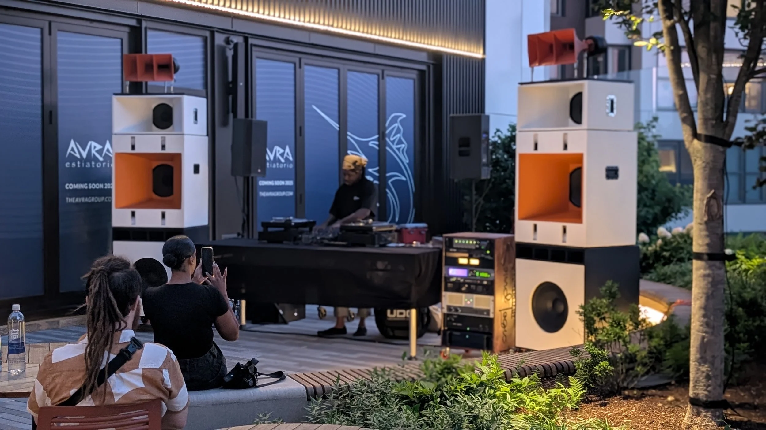 An outdoor music event with a DJ playing on a stage set up with large white speakers and orange accents, surrounded by people watching and taking photos, with trees and modern buildings in the background.