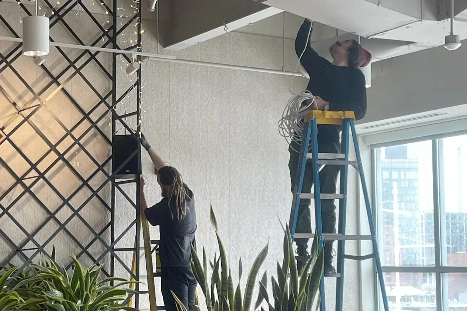 Two people installing or maintaining audio-visual equipment on a wall; one person standing on a ladder with a bundle of cables, another standing on a ladder reaching the ceiling.