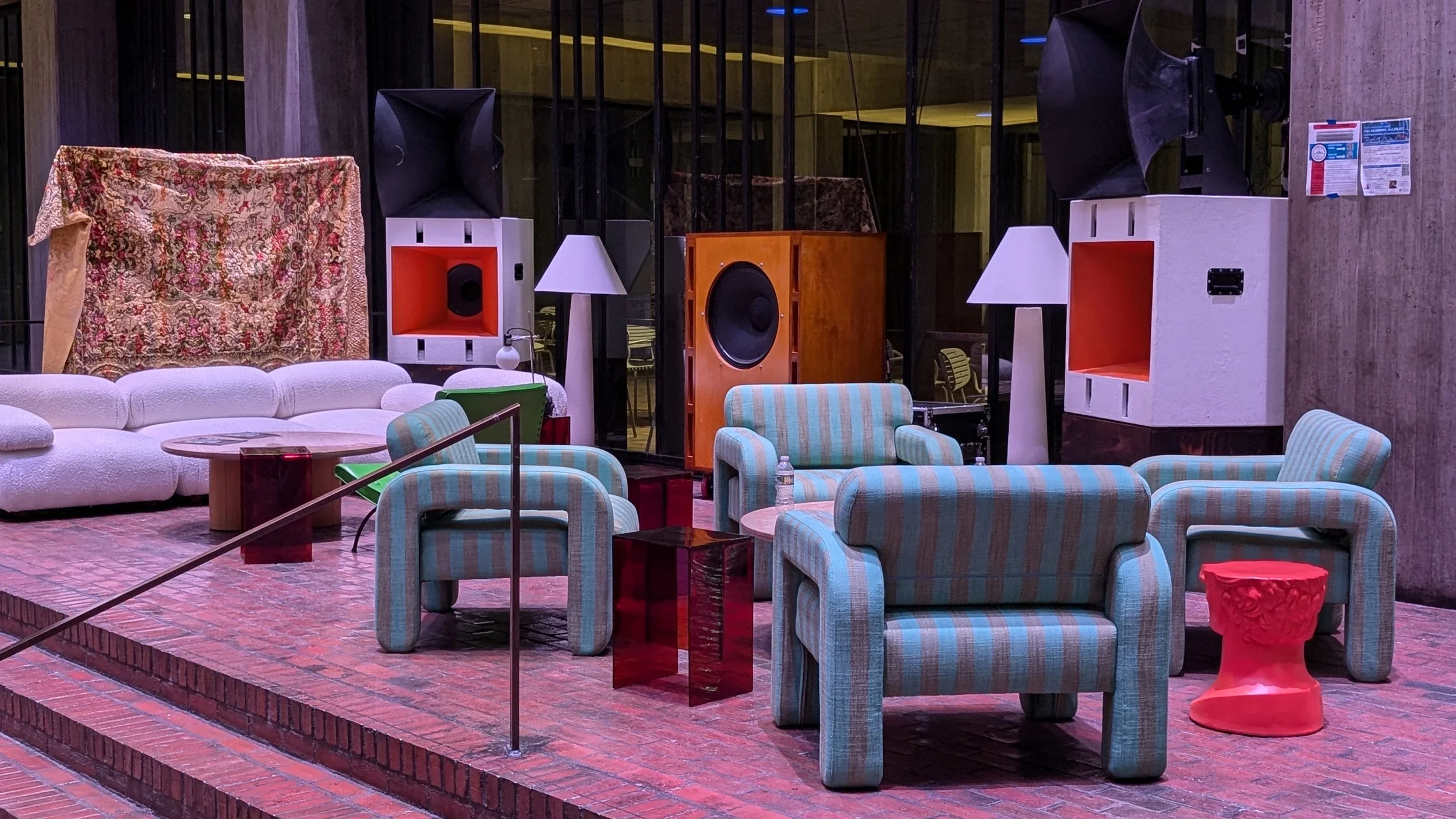 Cluttered indoor lounge area with mismatched furniture, large speakers, lamps, and a decorative tapestry in the background.