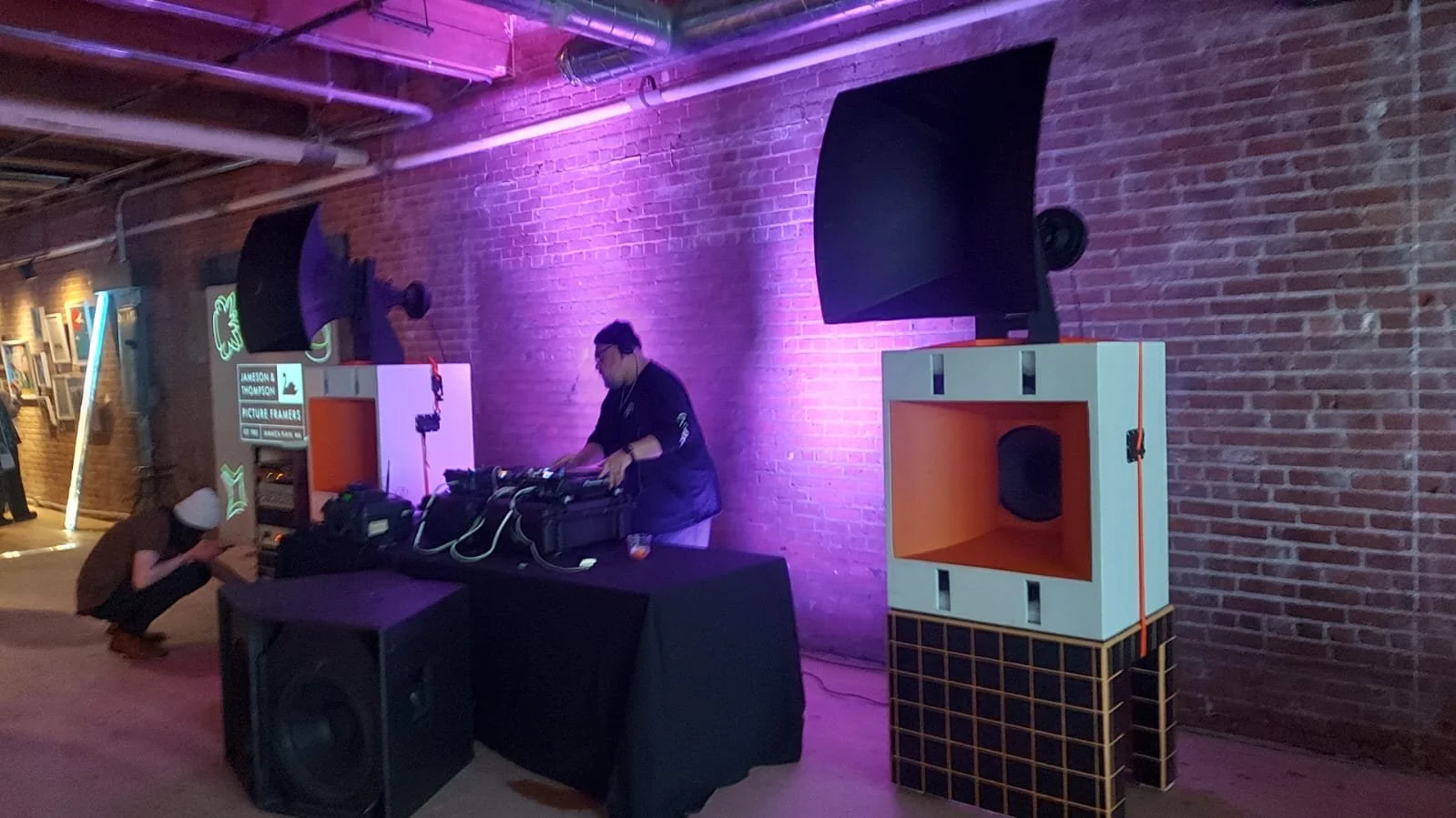 A DJ performing at an event with pink and purple lighting, with large black speakers and a woman crouching down nearby in front of a brick wall decorated with neon signs.