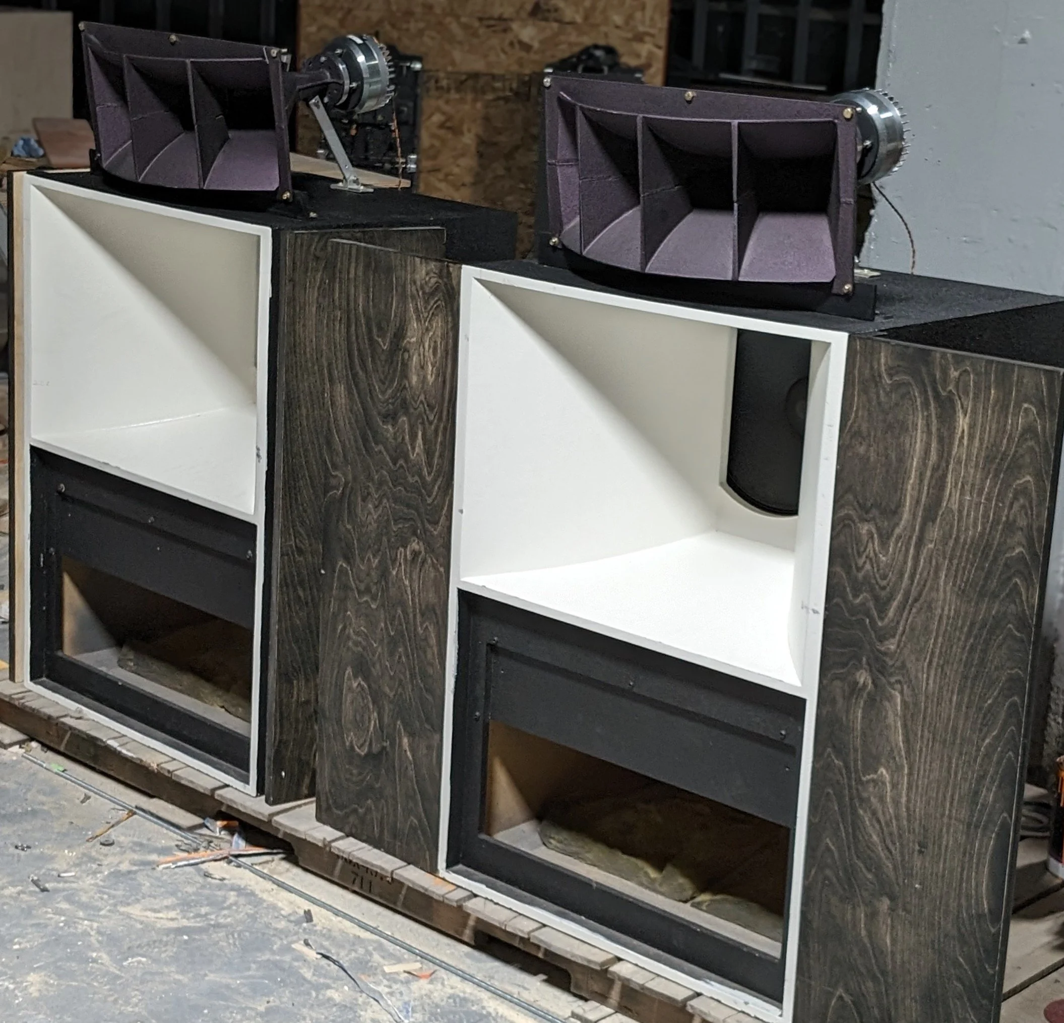 Two large speaker cabinets with open front panels and two smaller speaker units on top, in a workshop setting.