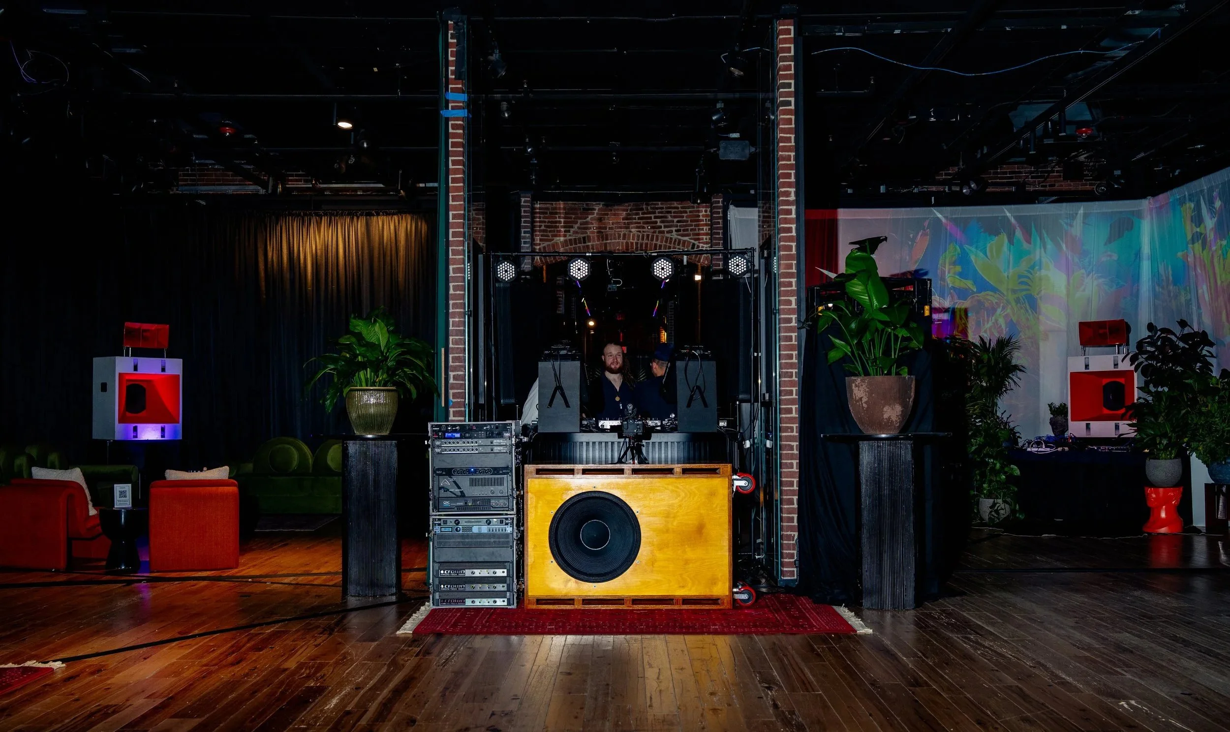 DJ setup with two people behind the turntables in a dimly lit venue, surrounded by plants, speakers, and seating area with couches and chairs.
