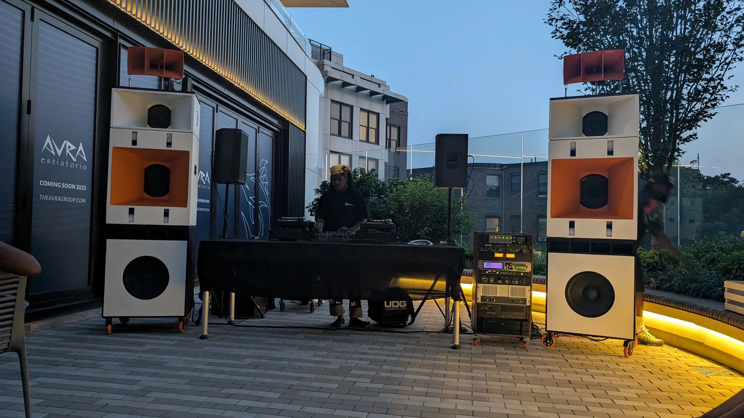 A DJ performing on stage with large speakers at an outdoor event during dusk, with a modern building and trees in the background.