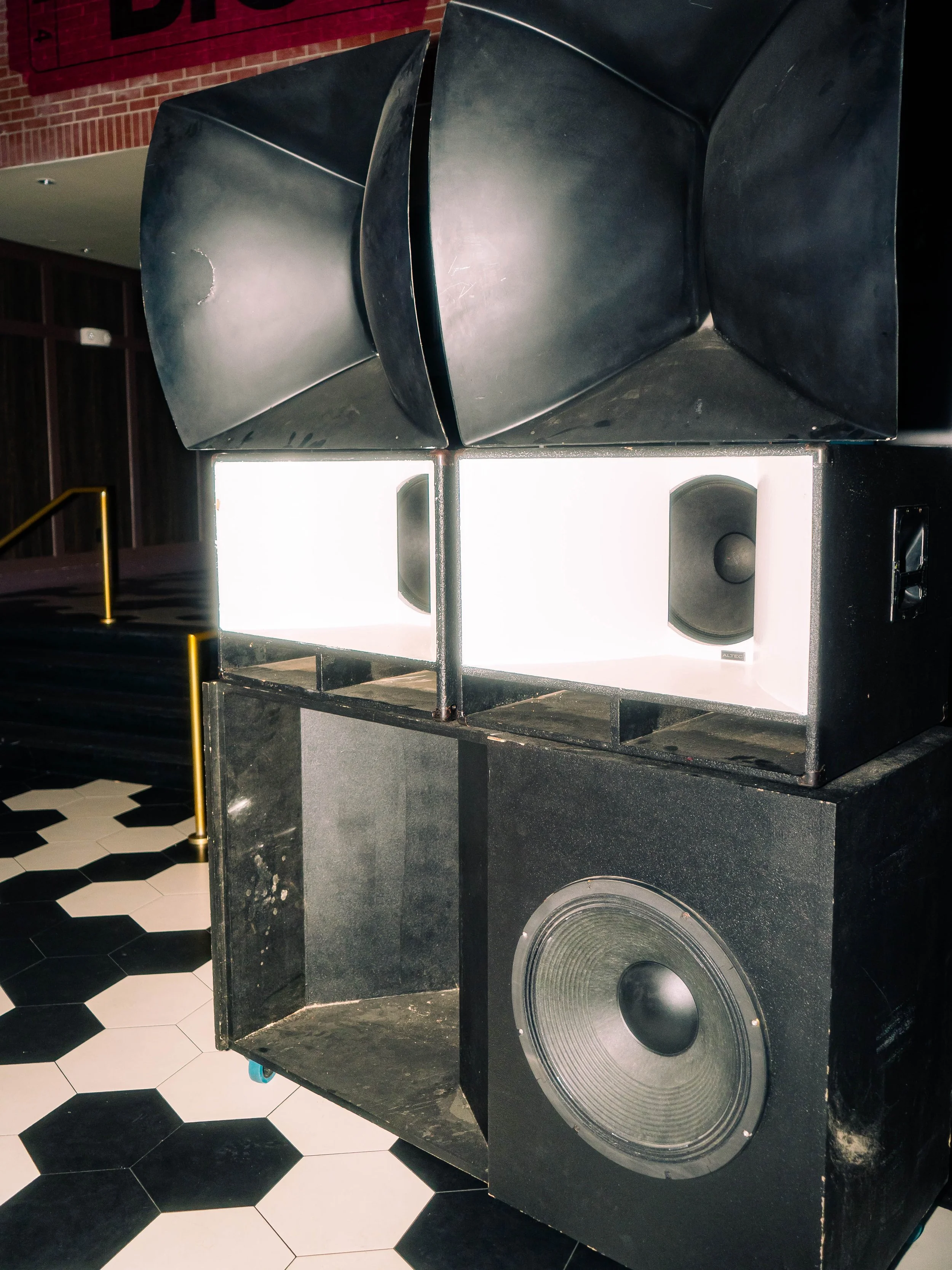 Several large black speaker cabinets stacked together with a bright white panel in the middle, with some speakers visible inside, on a black and white hexagonal tiled floor.