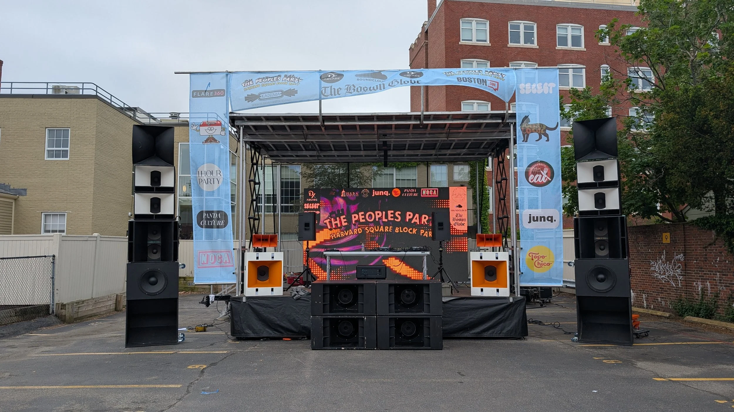 Outdoor stage setup with speakers and a digital screen displaying event information, banners with logos and illustrations, in a parking lot surrounded by buildings and trees.