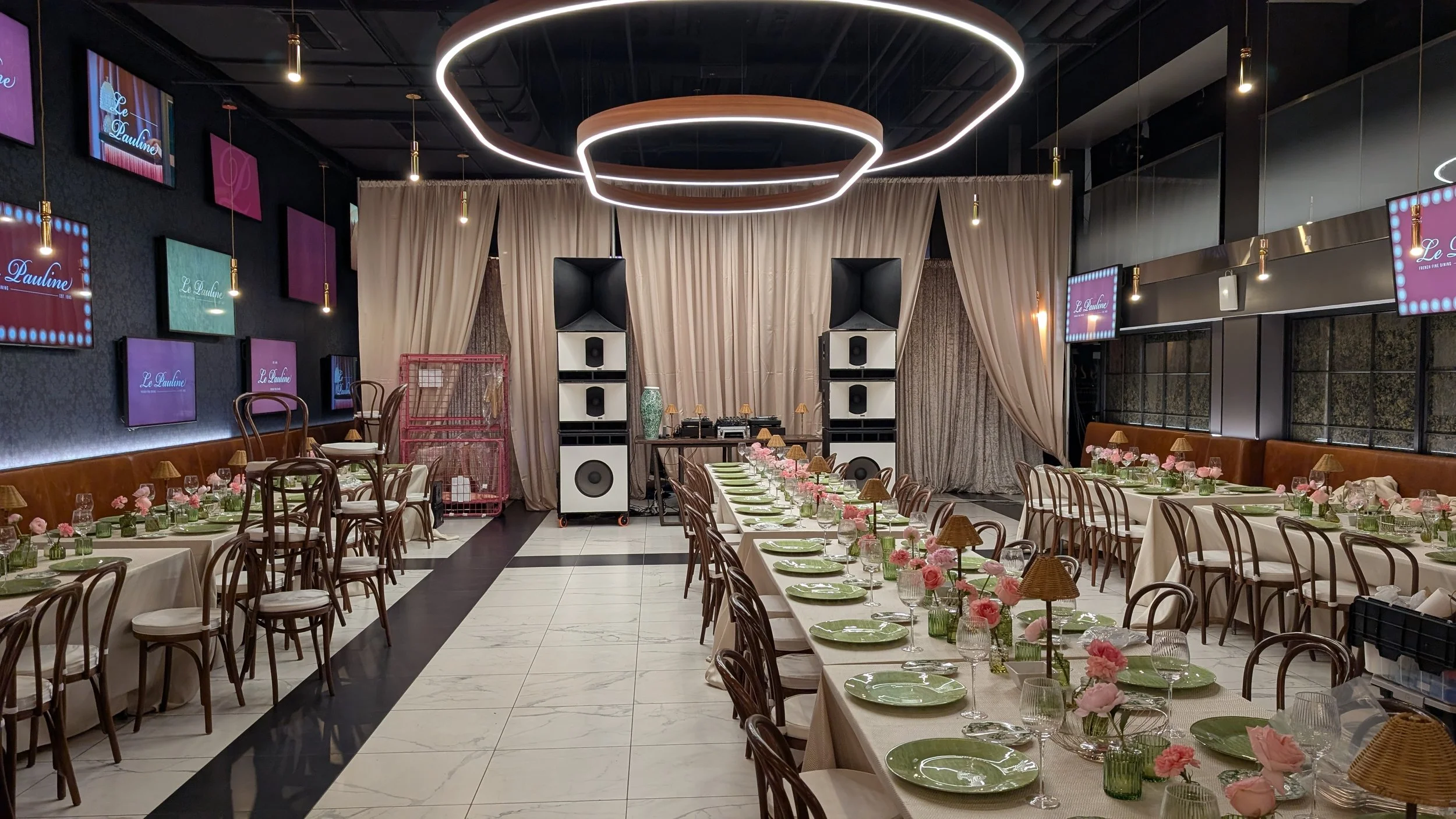 Set up for a celebration or event in a decorated indoor space with long tables, pink flowers, green plates, glassware, and small lamps, with a stage area and audio equipment at the front.
