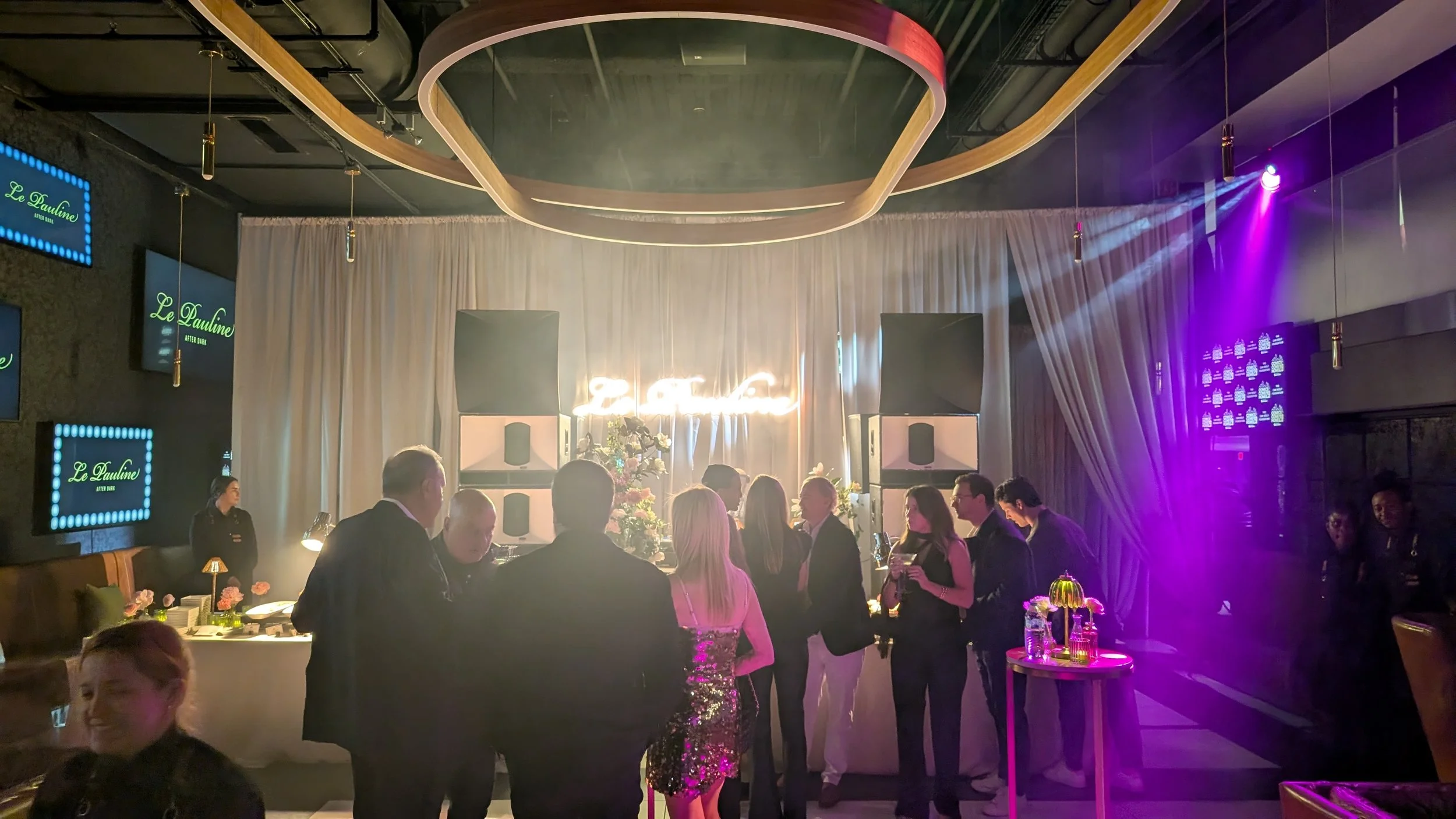 People gathered at a party or event in a decorated venue with a DJ or music setup, floral arrangements, and neon signs reading 'Le Pauline' and a neon sign that appears to read 'ZCTA'.