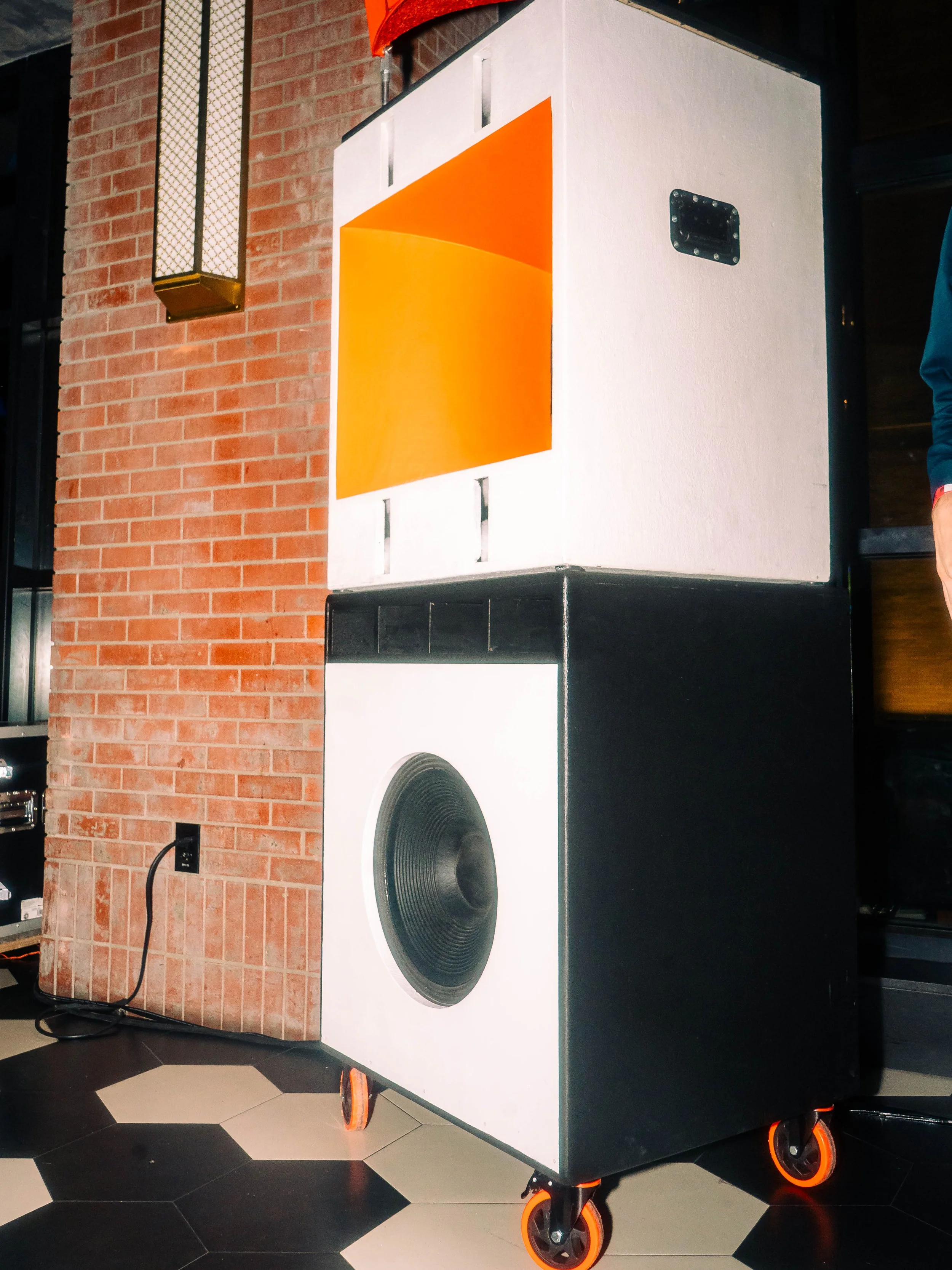 Large speaker with a black and white design, featuring a black speaker cone, on orange and black caster wheels, placed on a black and white tiled floor against a brick wall.