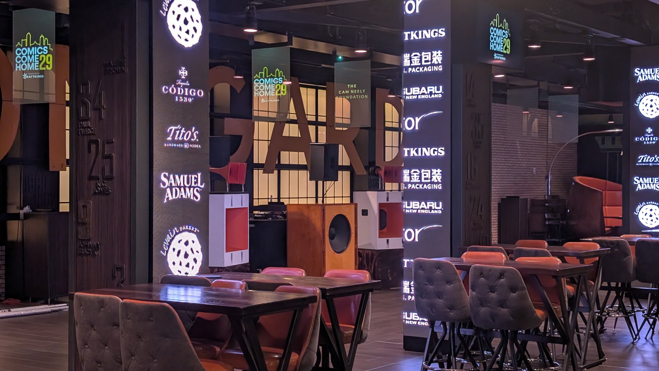 Indoor entertainment venue with tables and chairs, large illuminated screens displaying logos and text, including 'COMICS COME HOME 29', 'CODEG 1530', 'Tito's', 'SAMUEL ADAMS', and others, DJ equipment set up on stage, and decorative lighting.