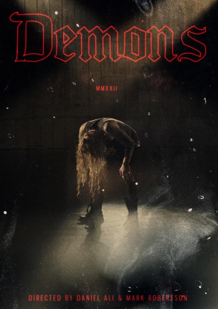 Poster for a show titled 'Demons' with a dark background, illuminated red neon-style text at the top, and an image of a woman with long hair bent over, wearing dark clothing, in the center. The bottom has red text stating 'Directed by Daniel Ali & Mark Robertson'.