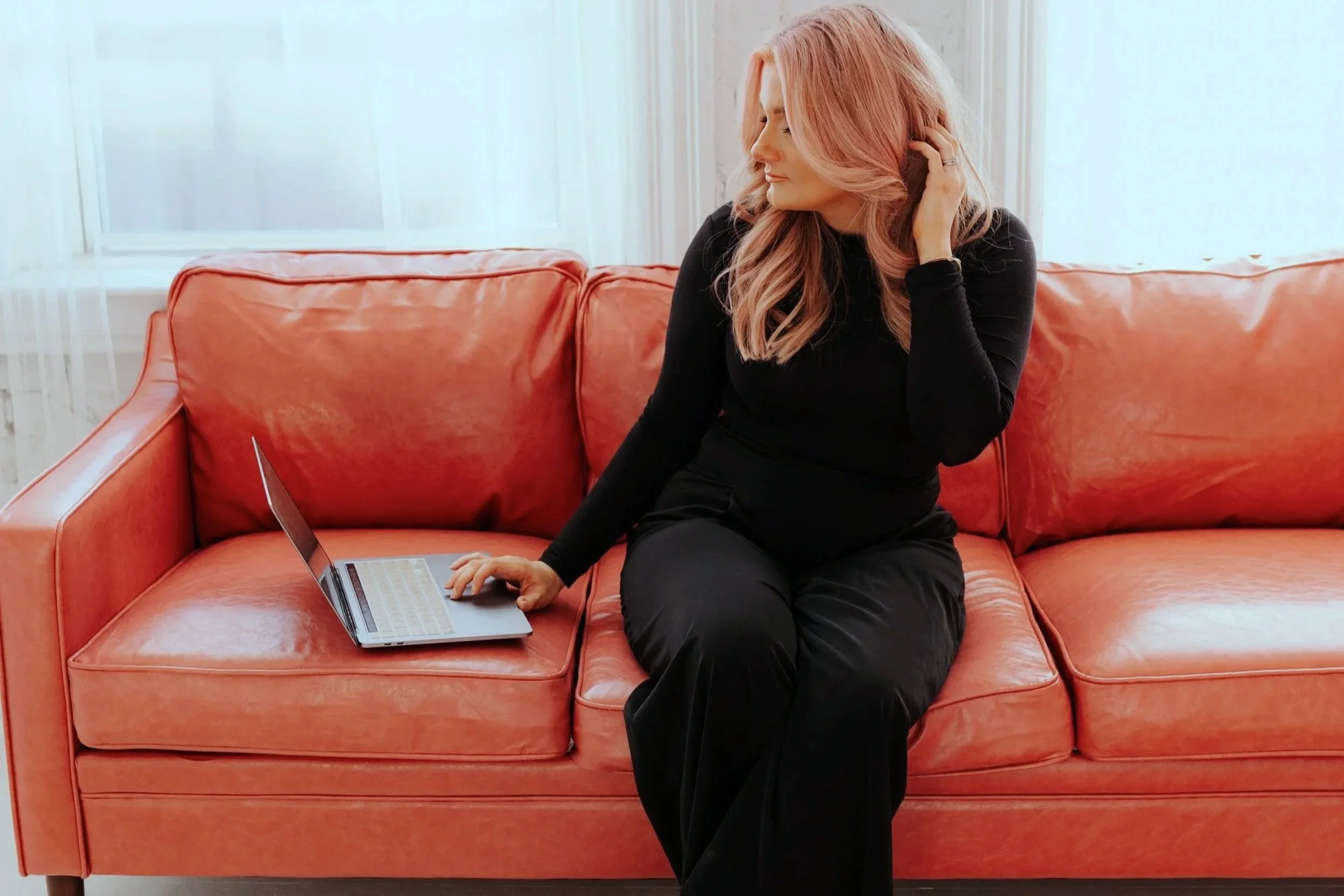 A woman with pink hair sitting on an orange sofa, looking down and to the side, touching her hair with one hand, with a laptop open on the sofa beside her.