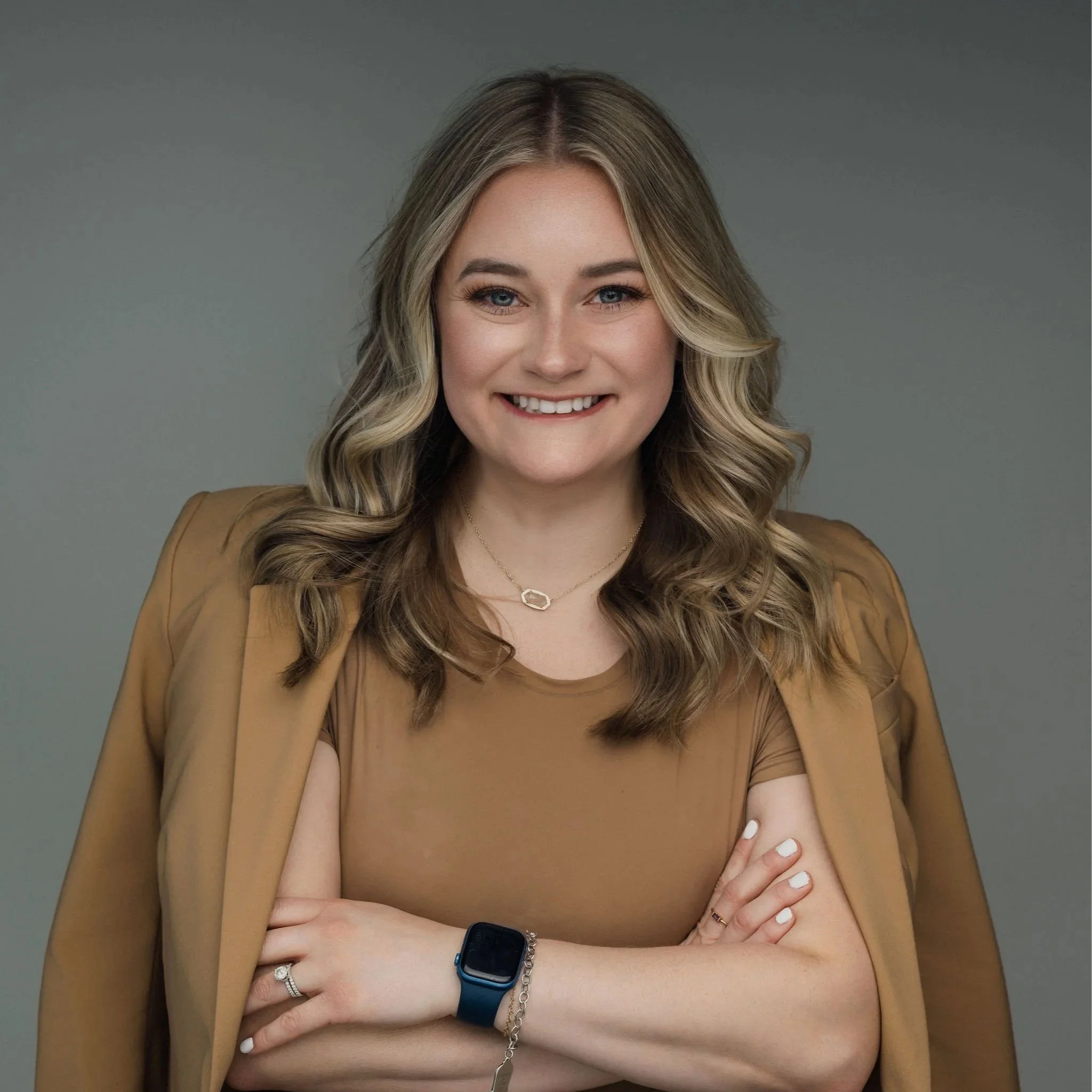 A smiling woman with wavy blonde hair, wearing a brown top and blazer, standing with her arms crossed, against a gray background.