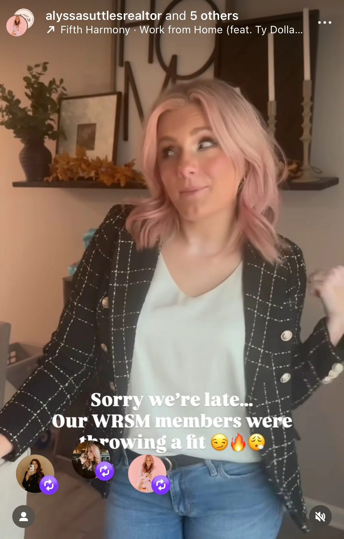 A woman with pink hair smiling and raising her fist, standing in a room with a shelf behind her decorated with framed pictures, a vase with greenery, and candles. She is wearing a black blazer with a white shirt and blue jeans.