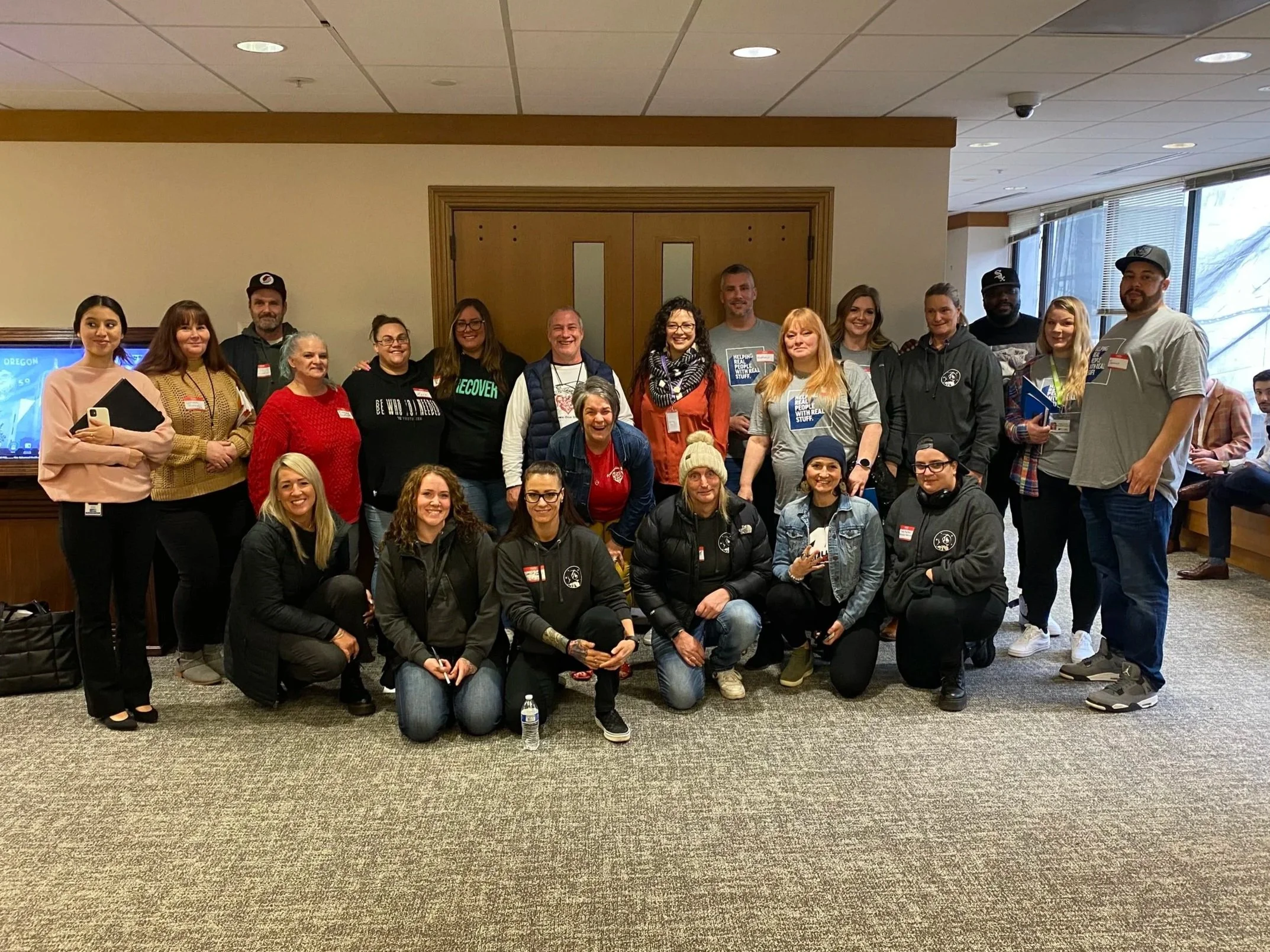 A group of diverse HJRA advocates pose for a photo indoors, standing and kneeling in front of wooden double doors, with windows on the right.