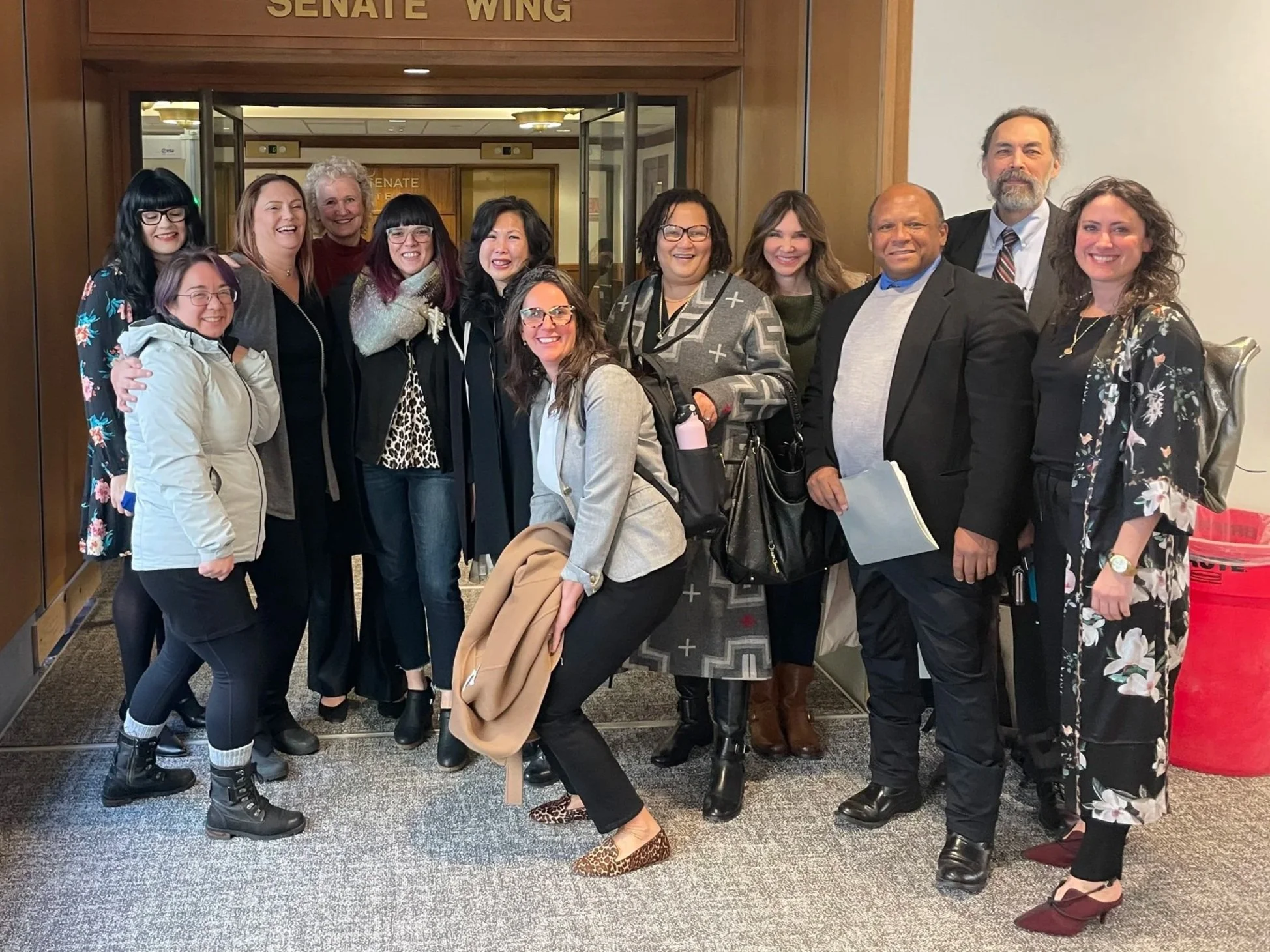 Group of diverse people posing for a photo outside a government building labeled 'Senate Wing'.