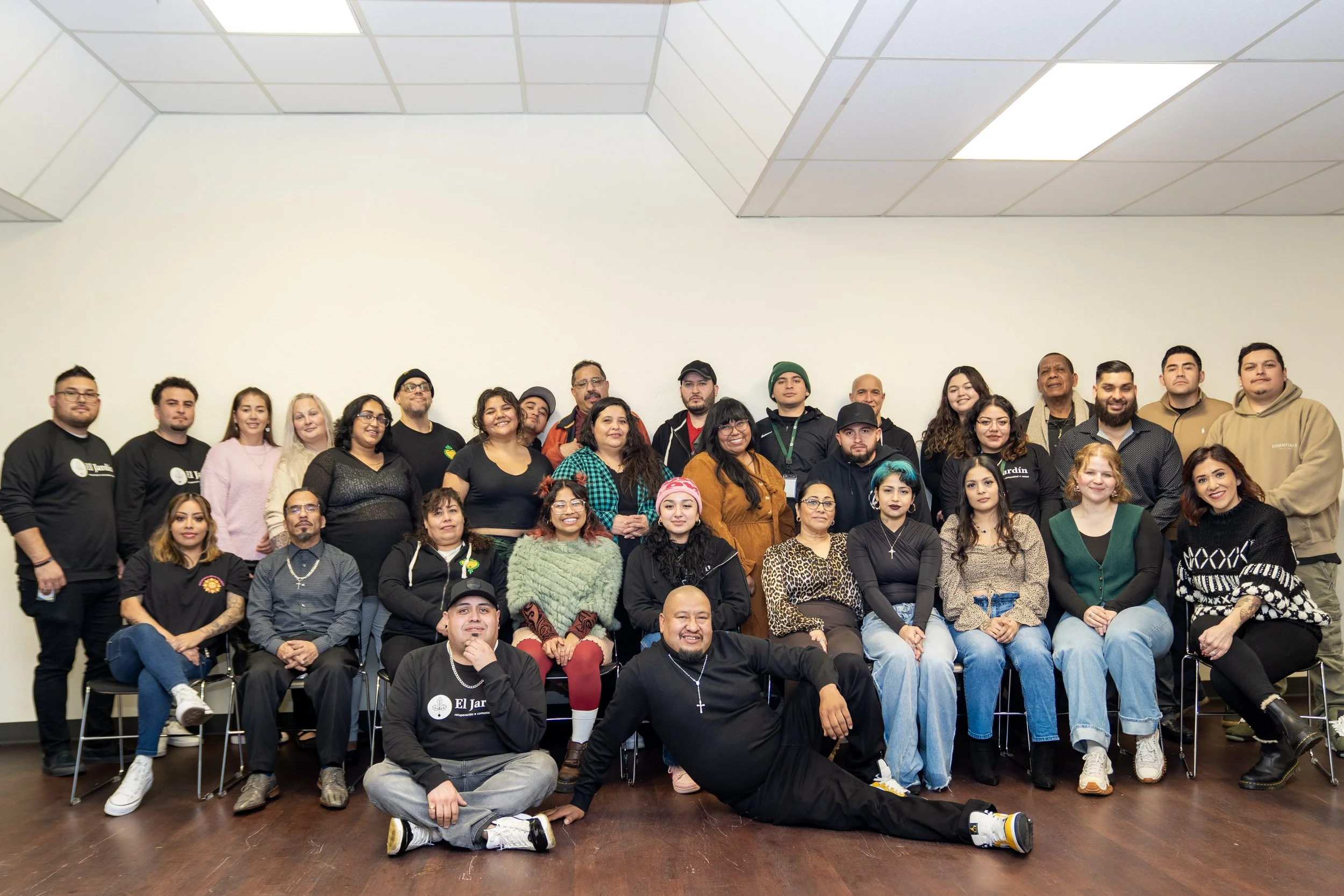 About 35 people who are part of El Jardín pose for a group photo in an indoor room with white walls and a wooden floor.