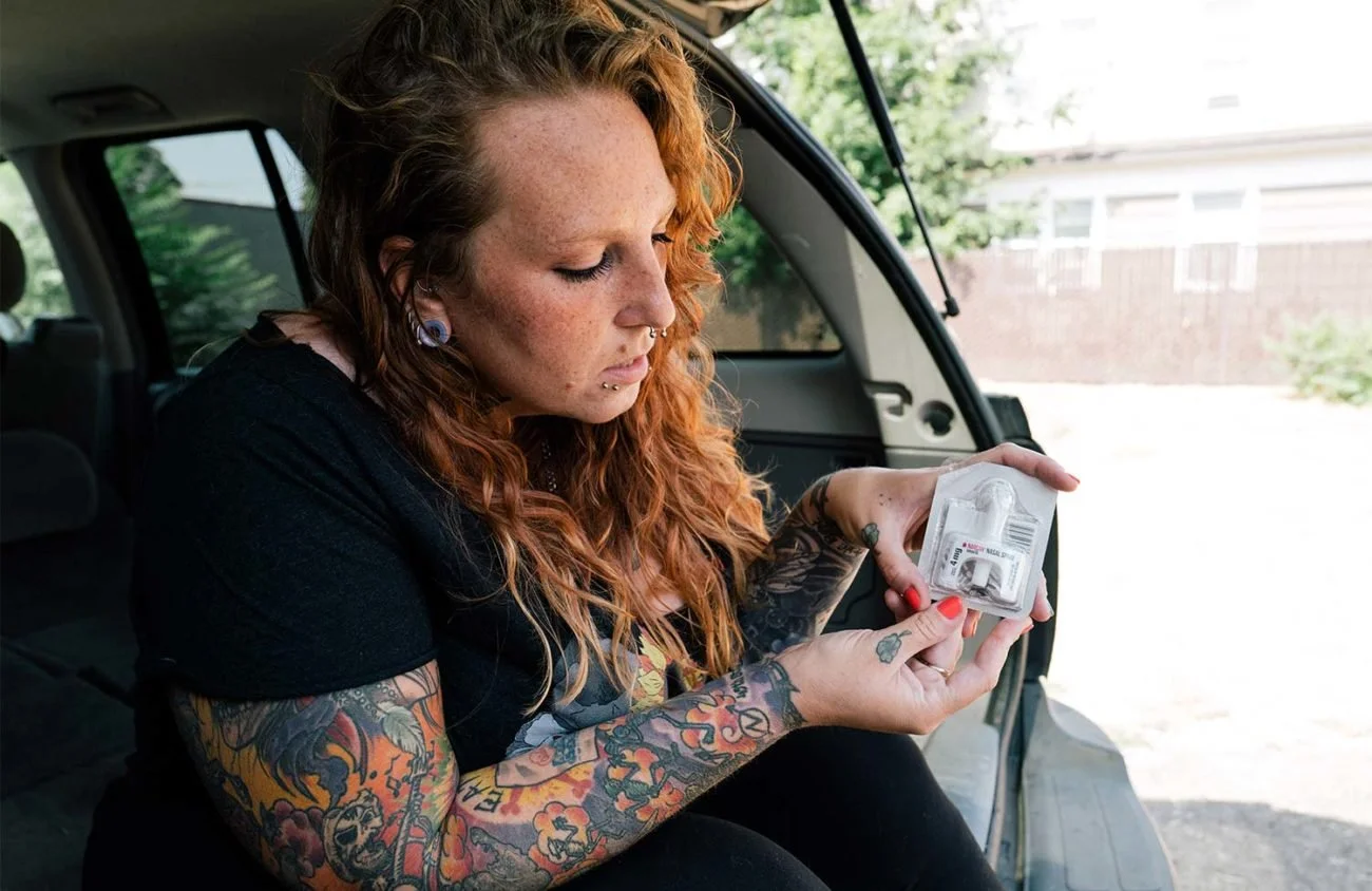 A woman from CORE with curly red hair, tattoos on her arms, and piercings, sits in a vehicle, looking at a package of supplies. 