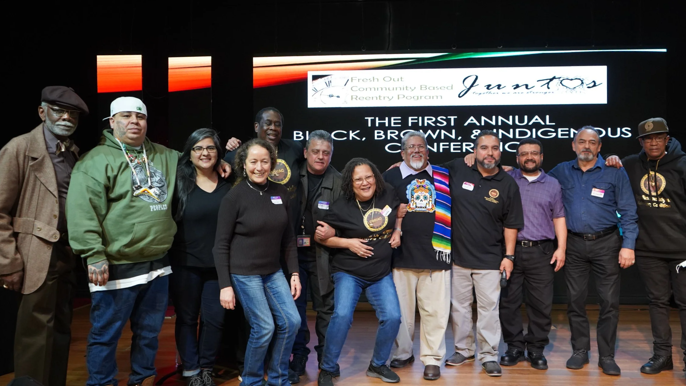 Diverse group of 12 people smile and pose in front of a sign saying "The First Annual Black, Brown, and Indigenous Conference".