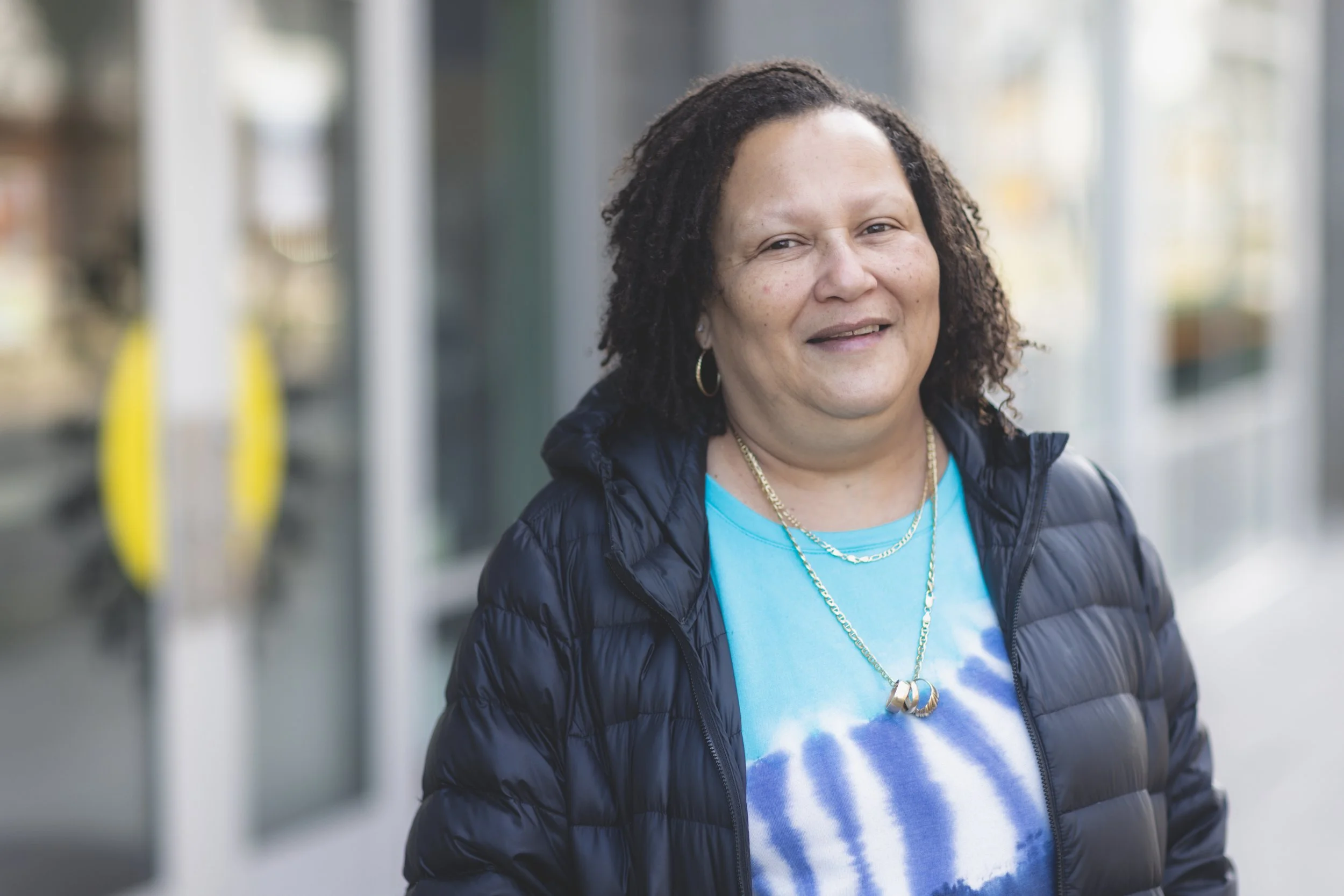 Executive DIrector of Miracles Club smiles. She wears a blue tie-dye shirt, a black puffy jacket, gold jewelry and is standing outdoors near a building.